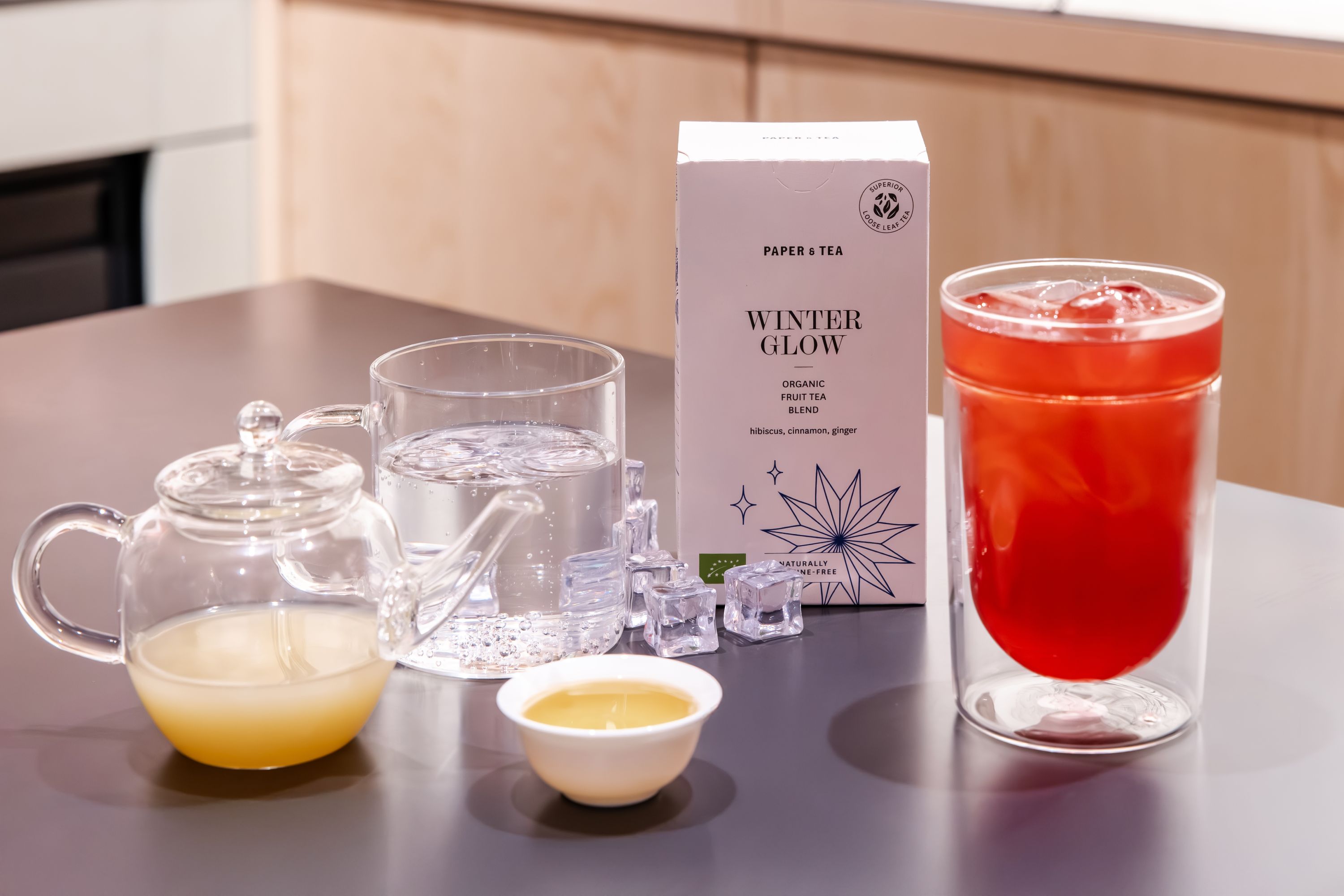 Weiße Verpackung von PAPER & TEA WINTER GLOW Bio-Früchtetee, roter Eistee in Glas und Tassen auf Tisch