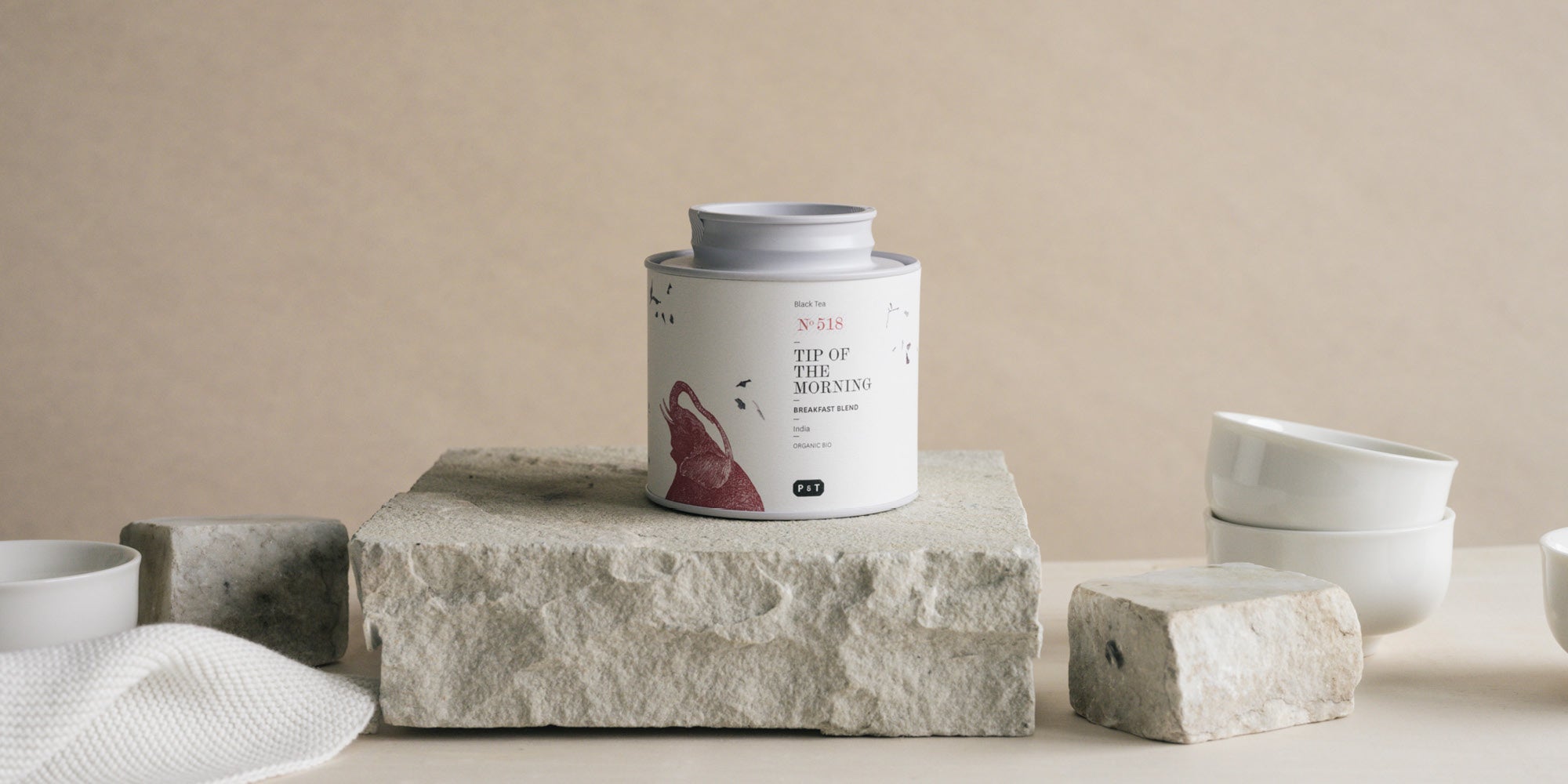 „Zylindrische weiße Teedose mit rotem Motiv und Schriftzug auf einem rechteckigen Steinblock; umgeben von kleinen weißen Schalen, weiteren Steinblöcken und einem Stück weißem Stoff vor beigefarbenem Hintergrund