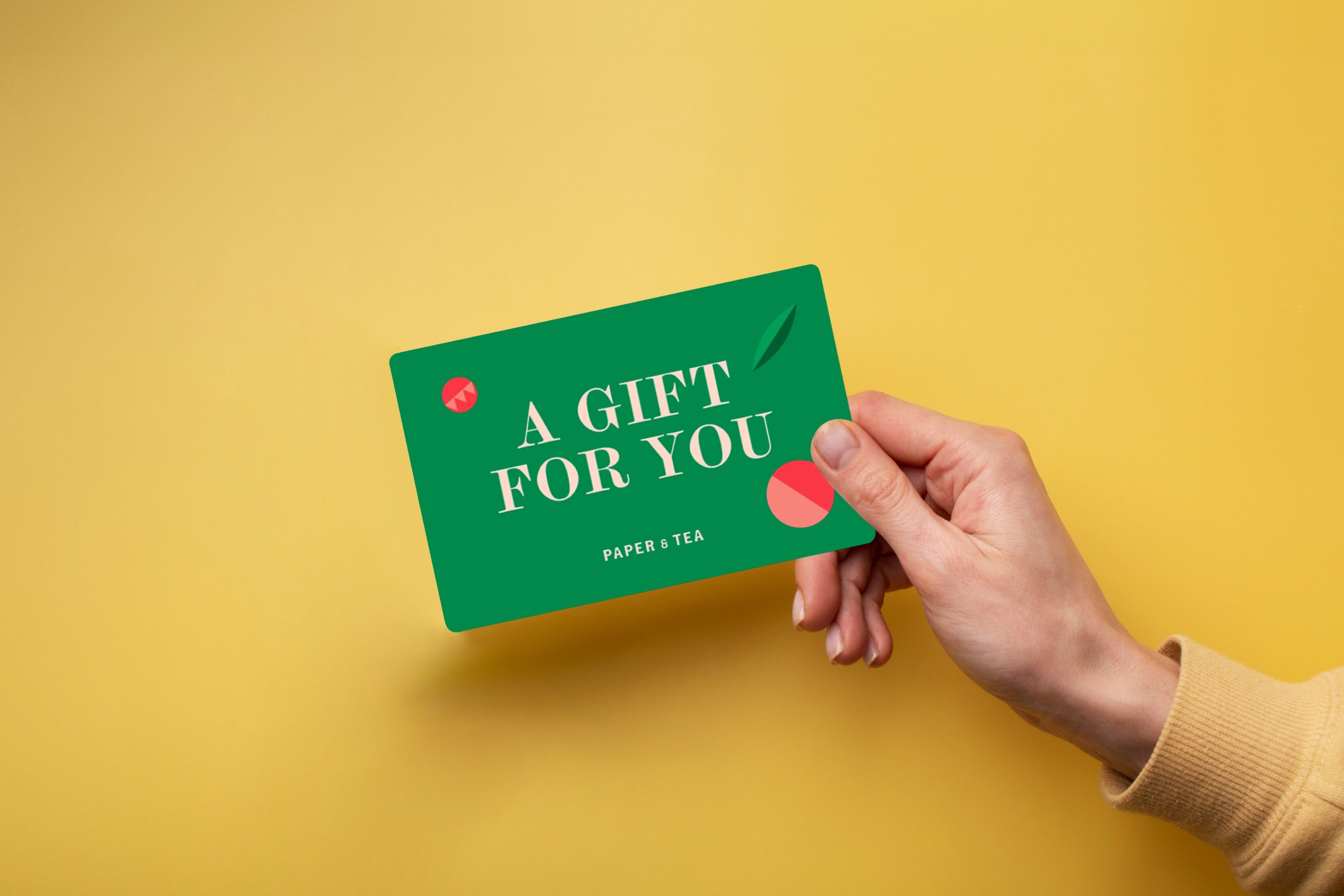 Hand hält eine grüne Geschenkkarte mit der Aufschrift ‚A GIFT FOR YOU‘ und ‚PAPER + TEA‘ vor gelbem Hintergrund