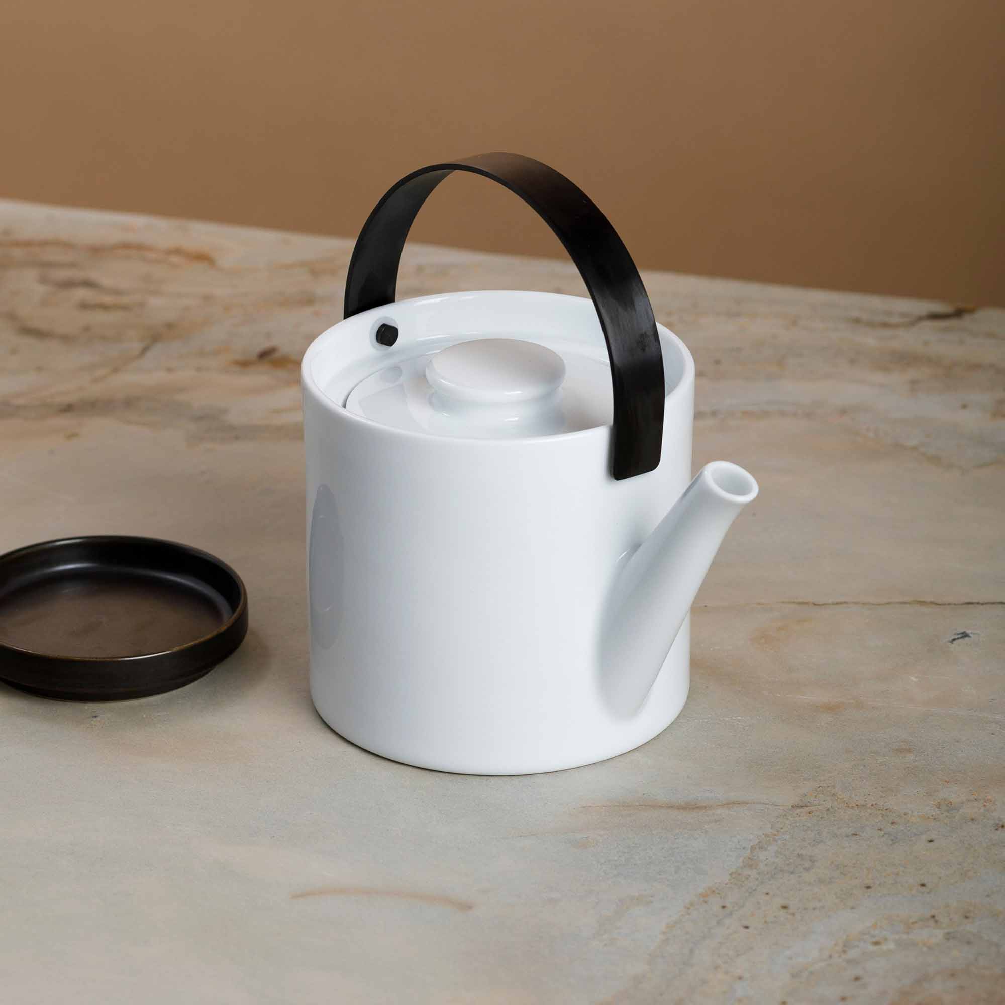 Minimalistische weiße Keramikteekanne mit schwarzem Henkel auf marmorierter Steinfläche, neben rundem Untersetzer.