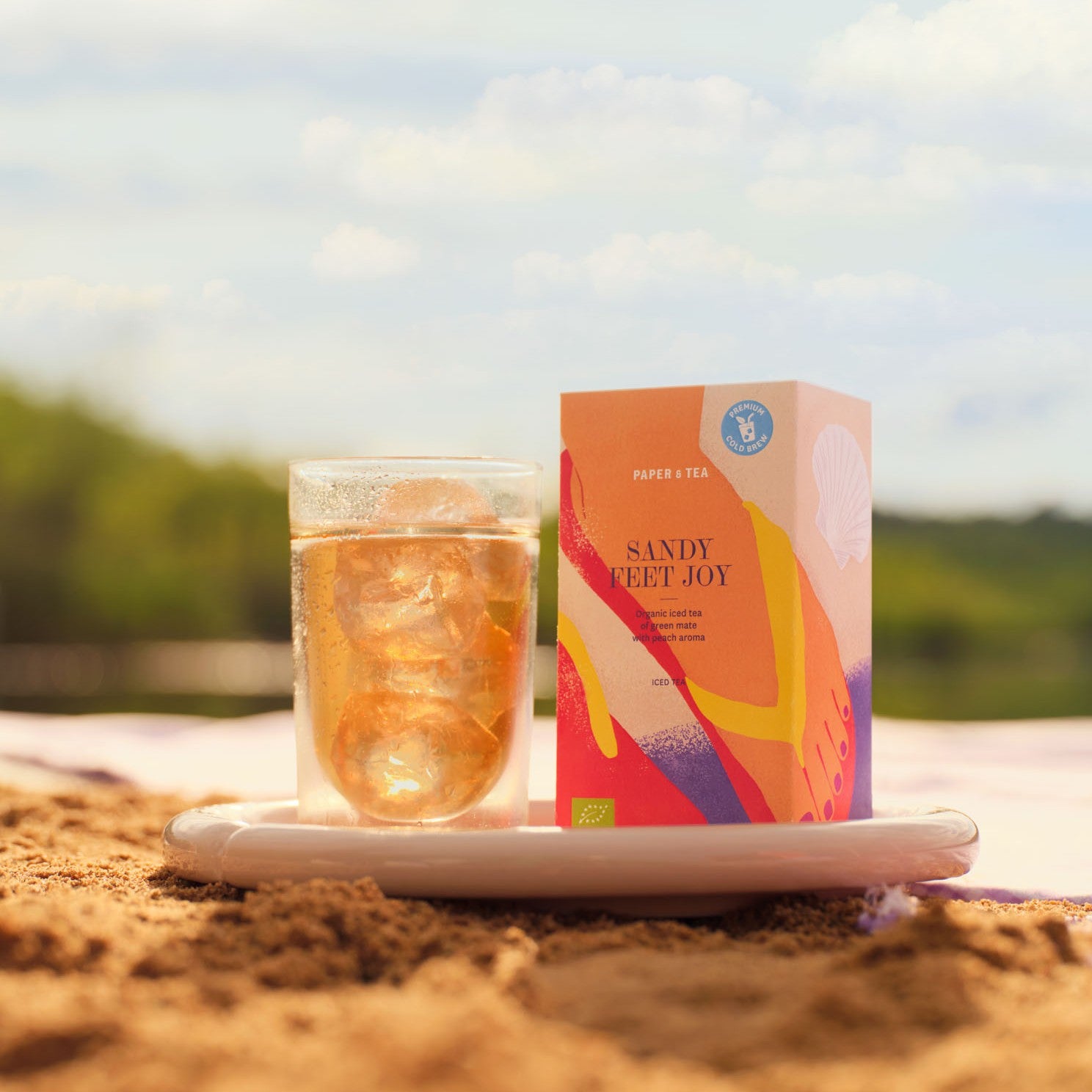 PAPER & TEA Sandy Feet Joy Bio-Eistee Box neben Glas mit Eiswürfeln auf Sandstrand