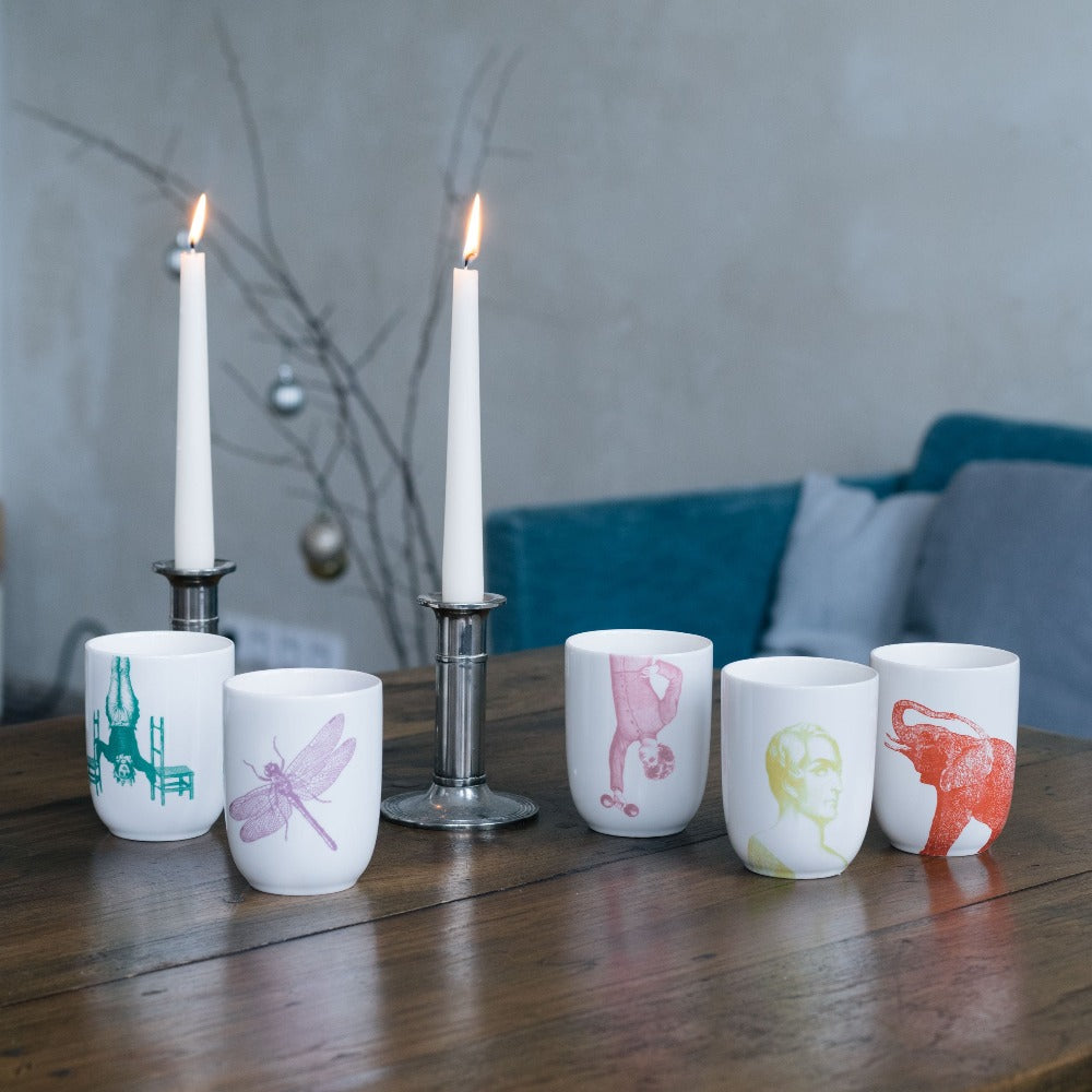 Holztisch mit zwei brennenden weißen Kerzen in silbernen Haltern und fünf Tassen mit bunten Illustrationen.