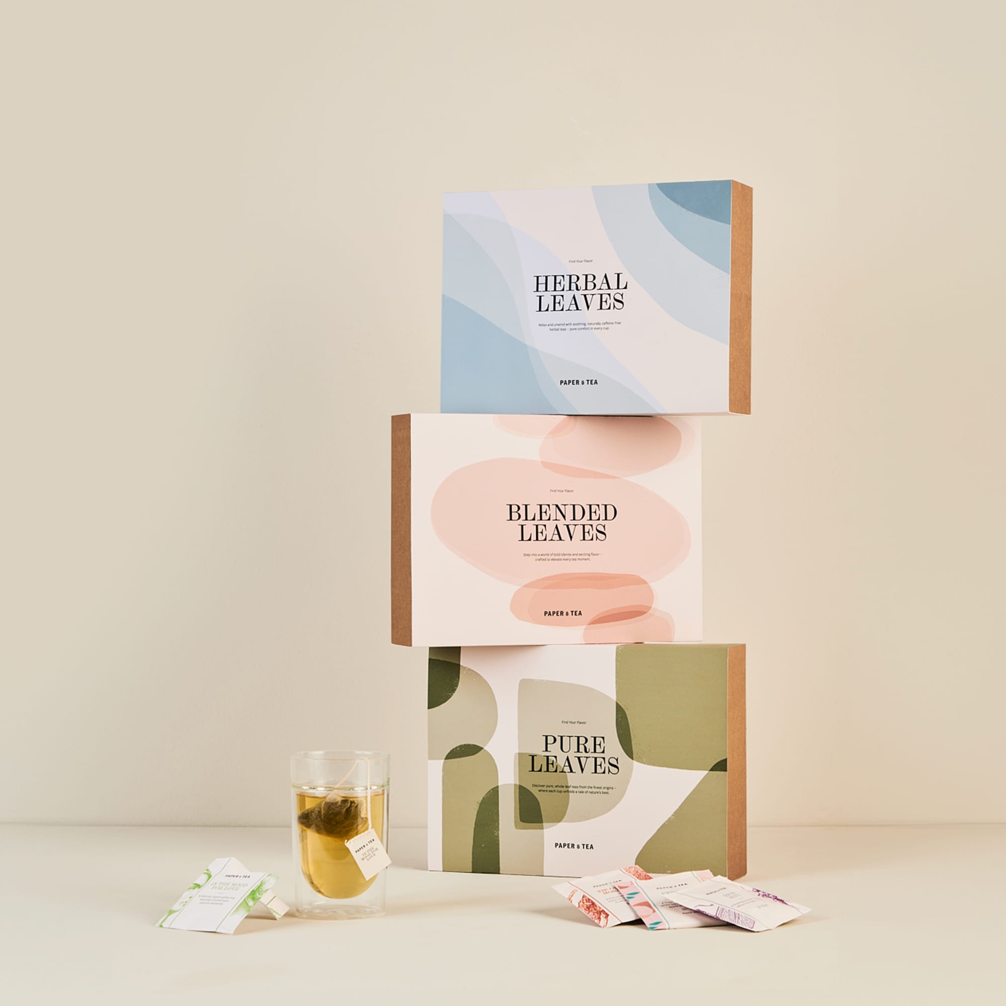 Drei pastellfarbene Paper & Tea Teeboxen, ein Glas Tee und verstreute Teebeutel auf neutralem Hintergrund.