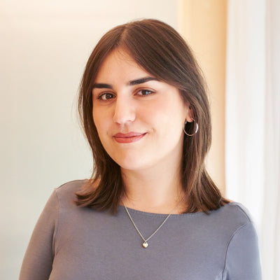 Porträt einer jungen Frau mit braunen Haaren, grauem Oberteil und silbernem Herzanhänger vor unscharfem Hintergrund.