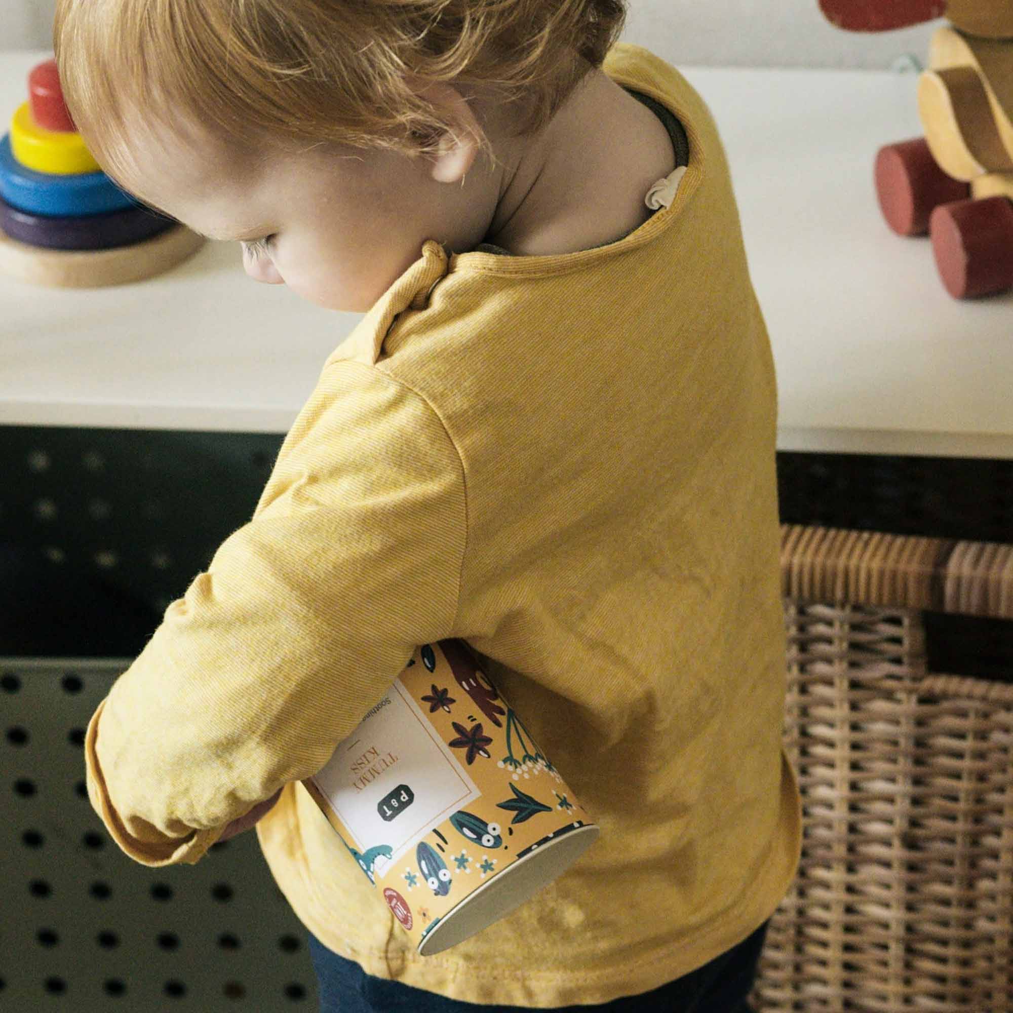 Kleinkind im gelben Shirt mit bunter P & T TUMMY KISS Dose in einem kinderfreundlichen Raum mit Holzspielzeug.