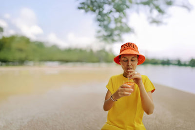 Frau in gelbem Kleid mit orangefarbener Mütze trinkt am Wasserufer in entspannter Atmosphäre