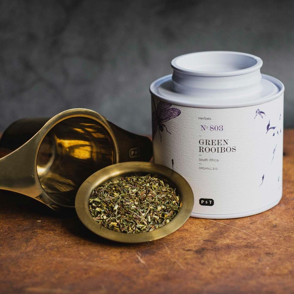 Weißer Tee-Behälter mit botanischen Illustrationen und grünem Rooibos-Tee in einem Messingsieb auf rustikalem Holz.