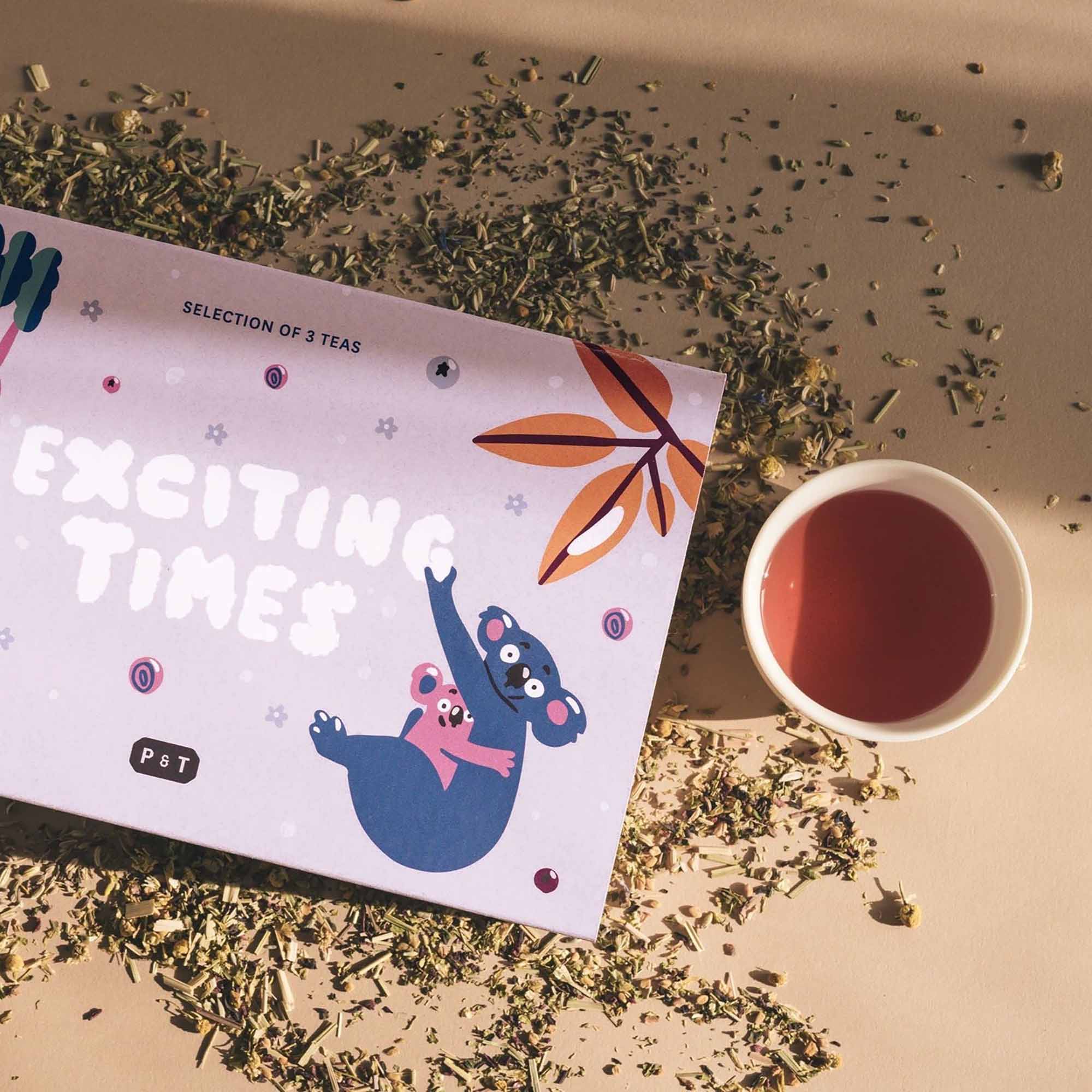 Rechteckige Lavendel-Teebox mit Koala-Illustrationen, drei Teesorten und weiße Tasse rötlichem Tee auf Beige.