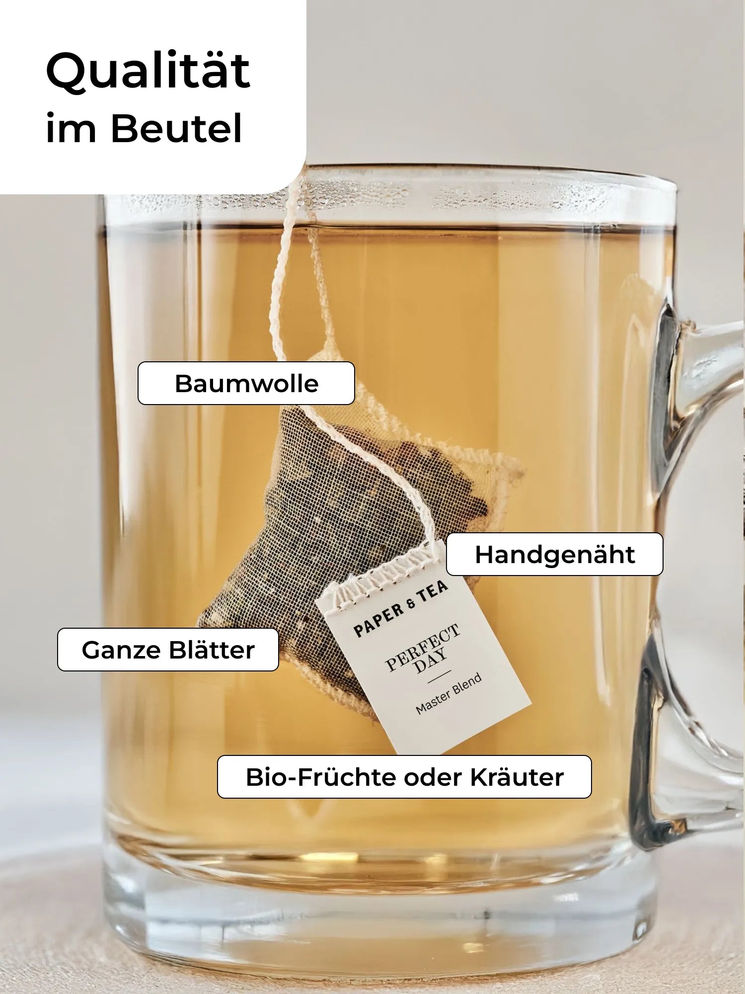 Gläserner Becher mit hellem Bernsteintee und Baumwoll-Teebeutel der Bio-Mischung von PAPER & TEA.