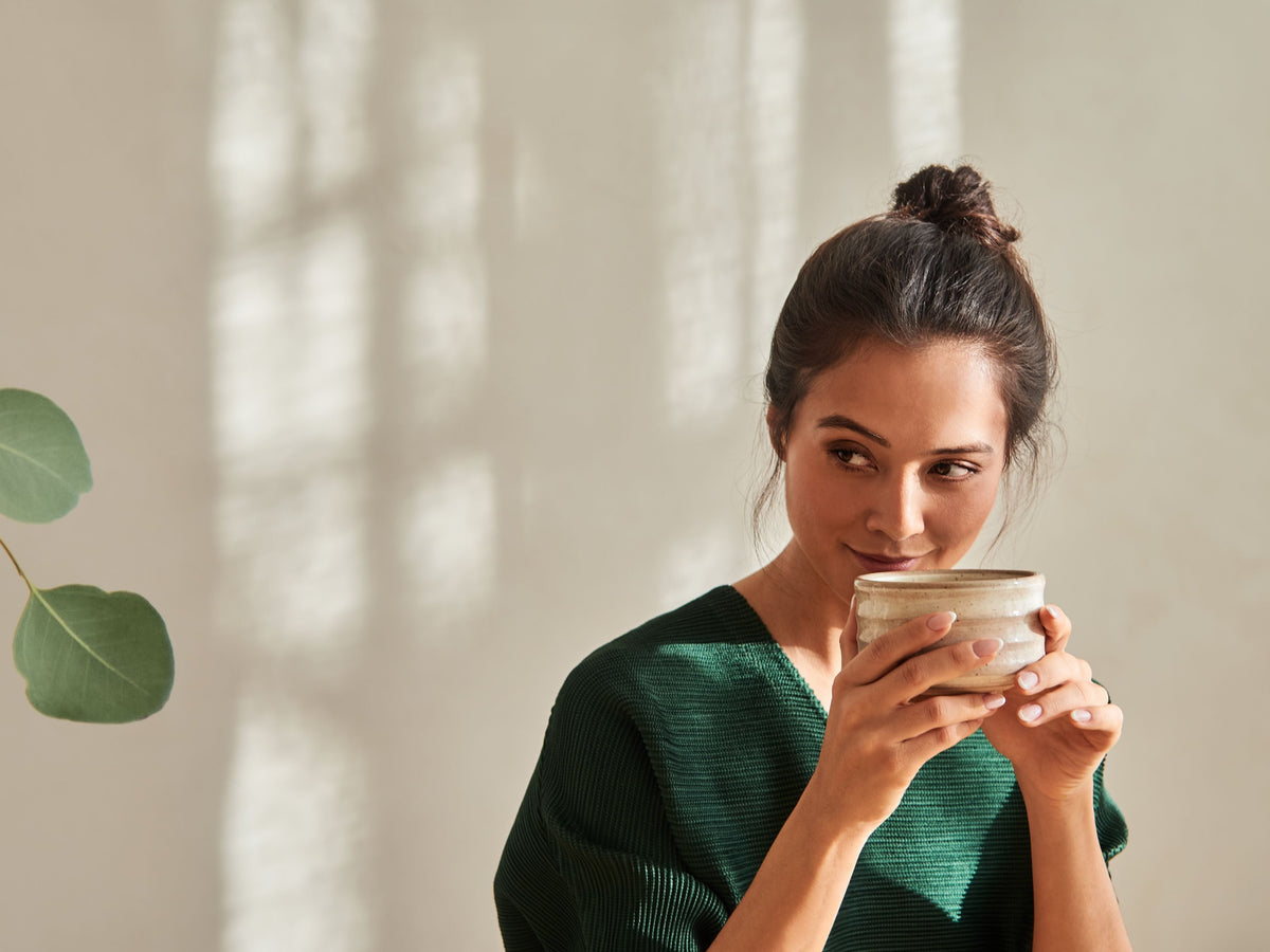 Frau in dunkelgrünem Pullover hält eine beige Keramiktasse vor ihrem Gesicht und lächelt sanft.