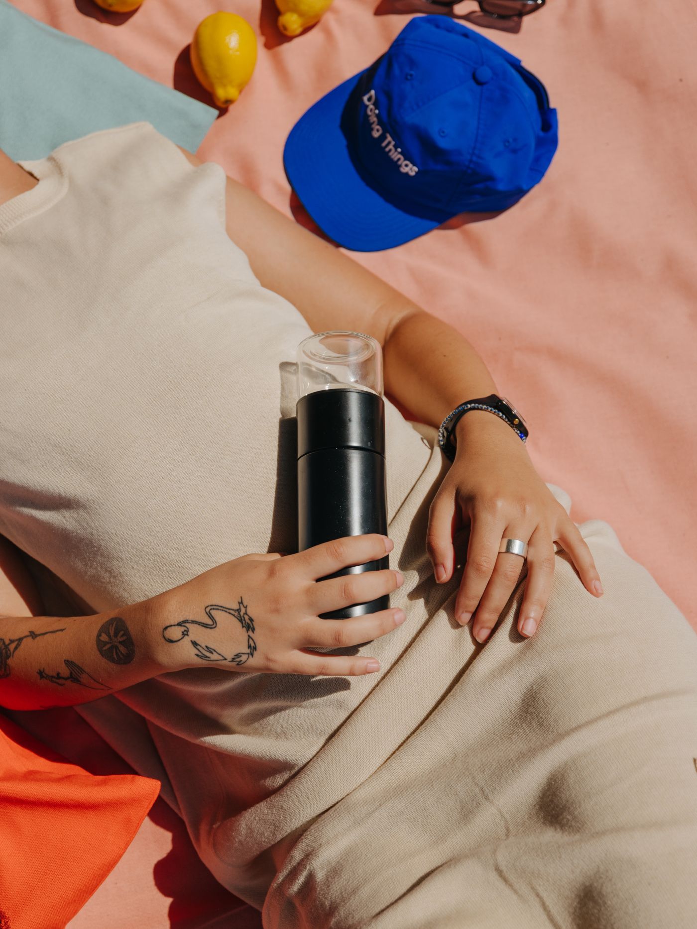 Person in beigem Kleid liegt auf buntem Stoff, hält schwarze Kaffeemühle, blaue Mütze mit 'Doing Things', Zitronen.