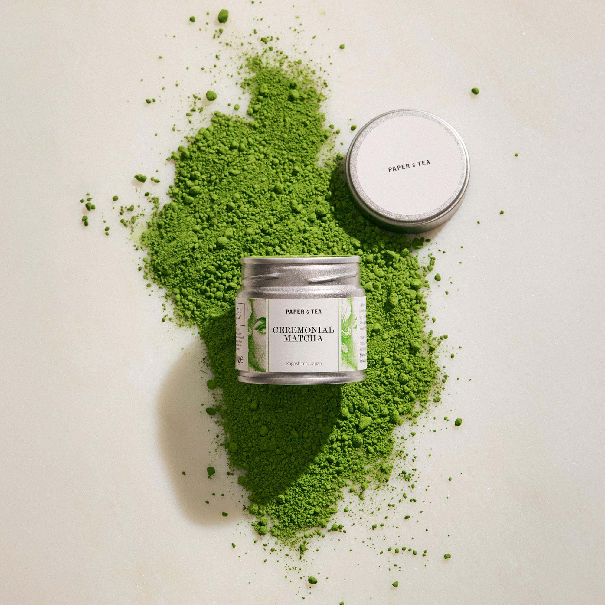 Kleine metallene Paper & Tea Matcha-Dose auf verstreutem, leuchtend grünem Matcha-Pulver aus Kagoshima, Japan