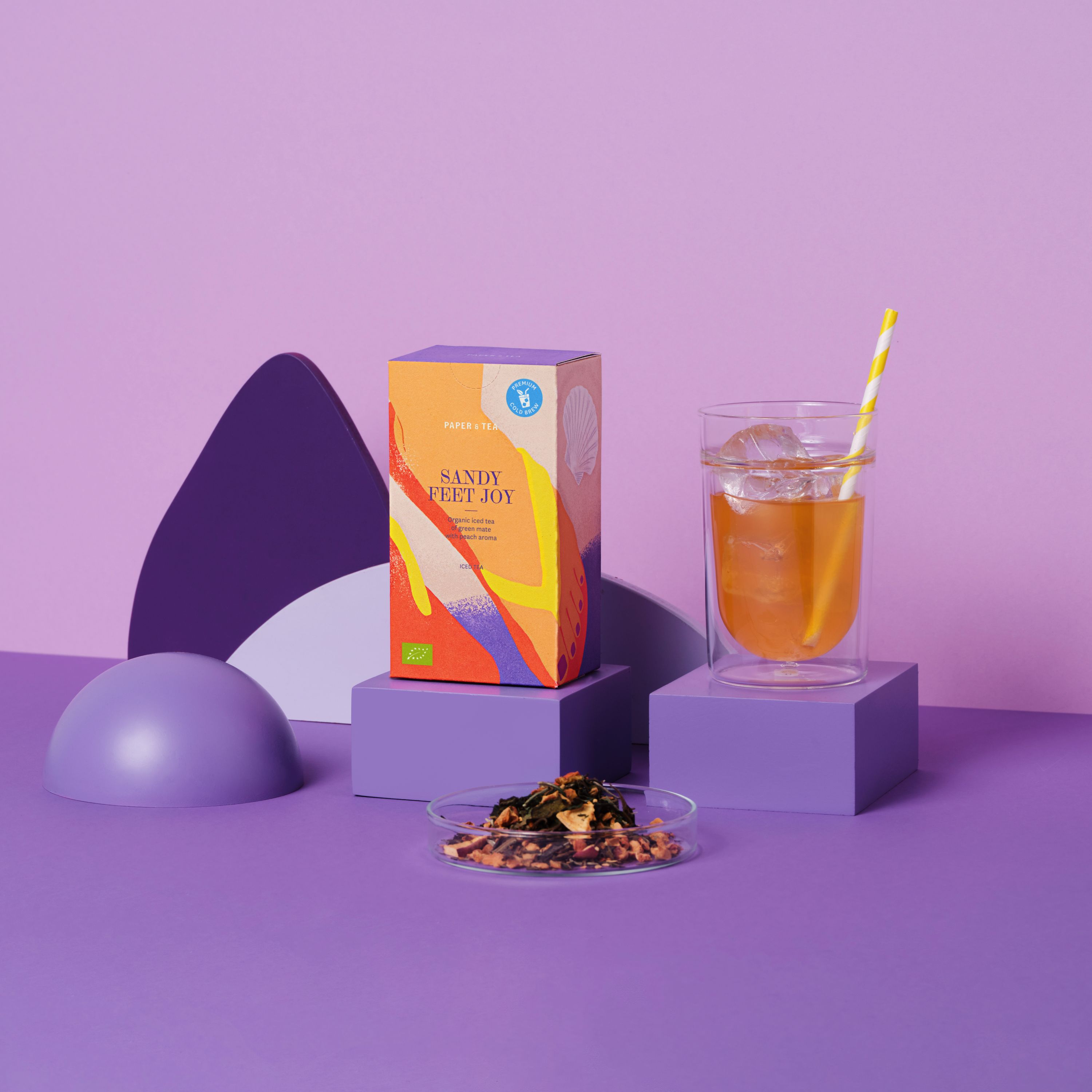 Bunte Verpackung von PAPER & TEA Iced Tea 'Sandy Feet Joy' mit Glas Eis, Strohhalm und losem Tee
