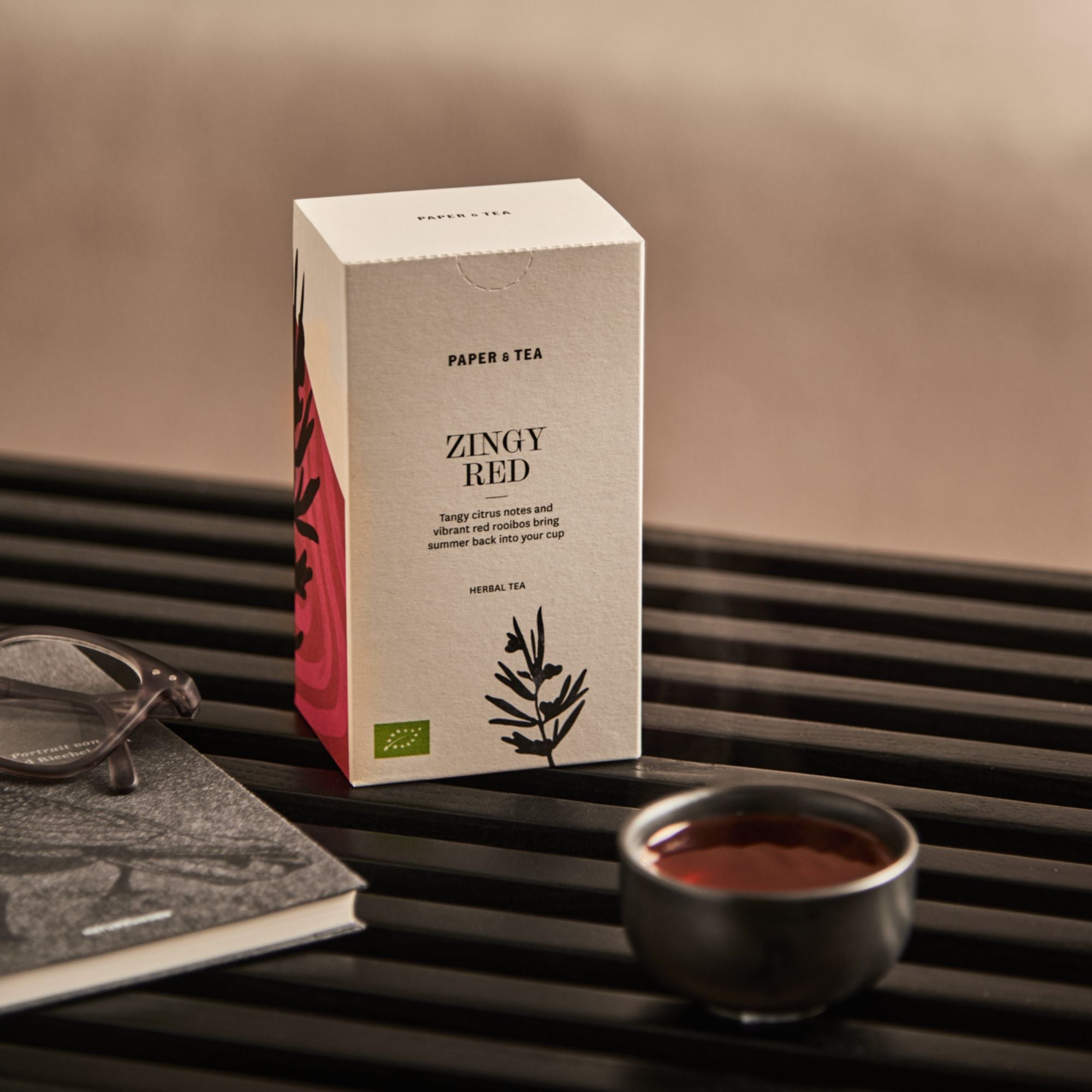 Paper & Tea Zingy Red Kräutertee-Verpackung neben roter Rooibos-Tasse, Buch und Brille auf dunklem Holz.