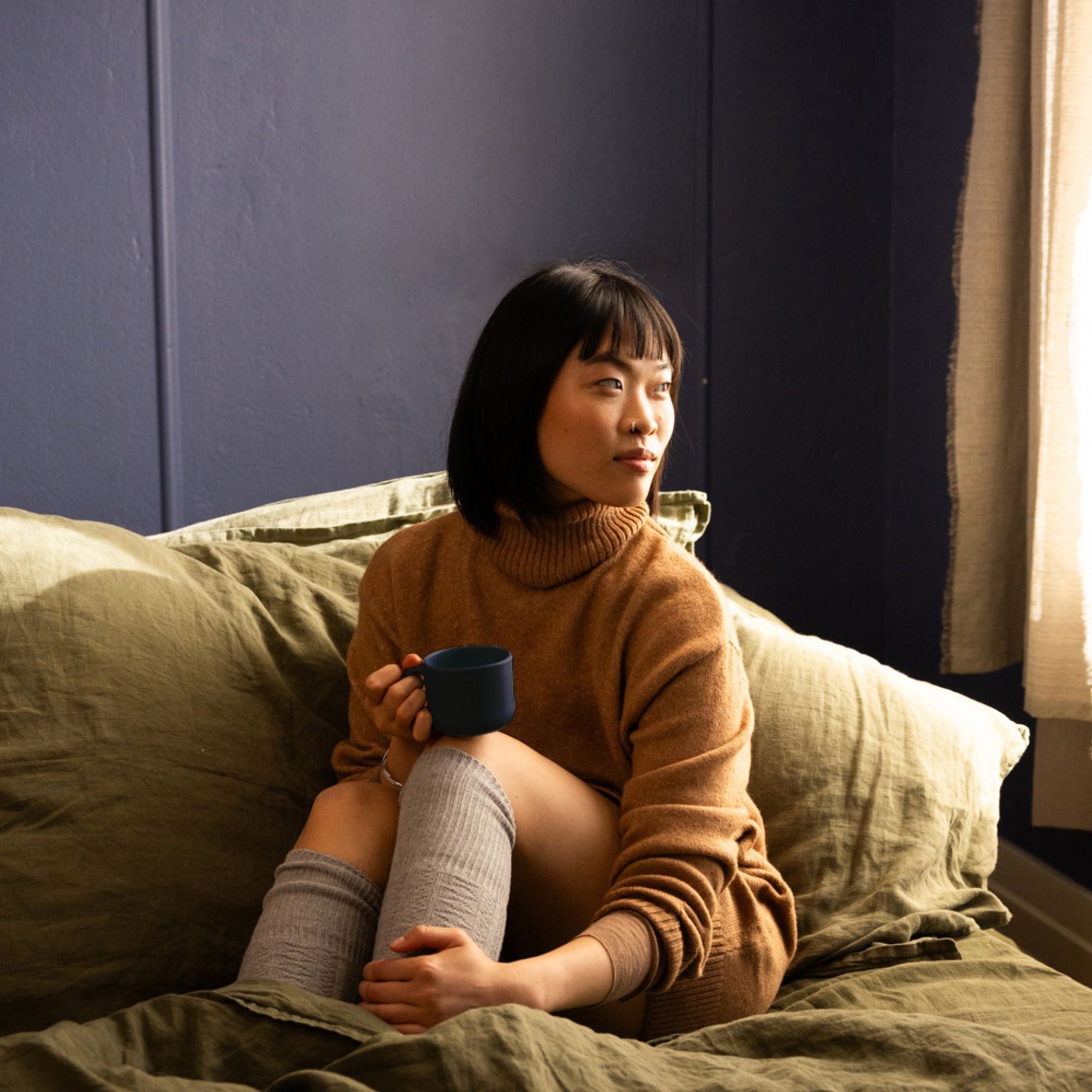 Junge Frau in braunem Rollkragenpullover sitzt auf Bett mit olivgrüner Bettwäsche, hält blaue Tasse