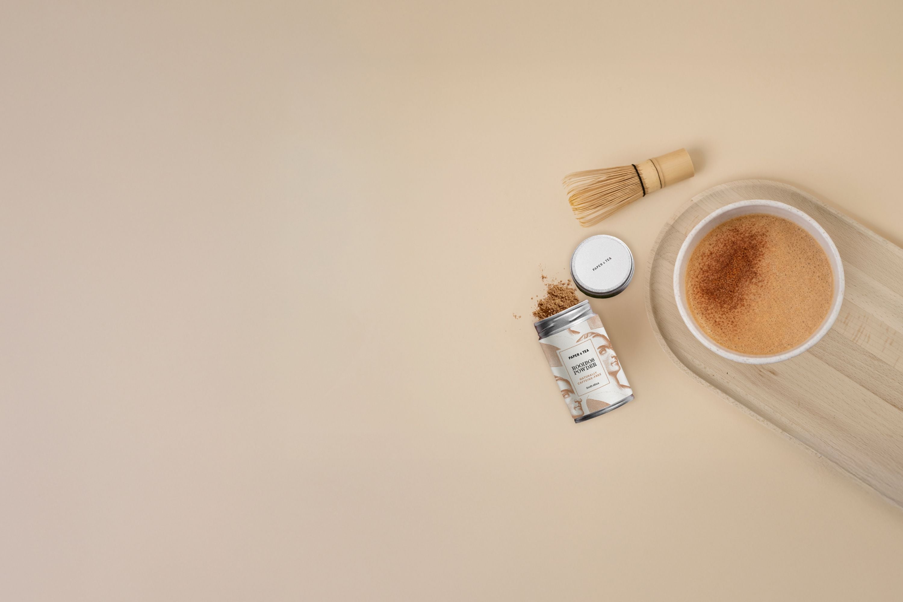 Offene Metallbüchse mit P&T Rooibos-Pulver, Bambusbesen und P&T Rooibos-Tee im Pappbecher auf Holztablett.