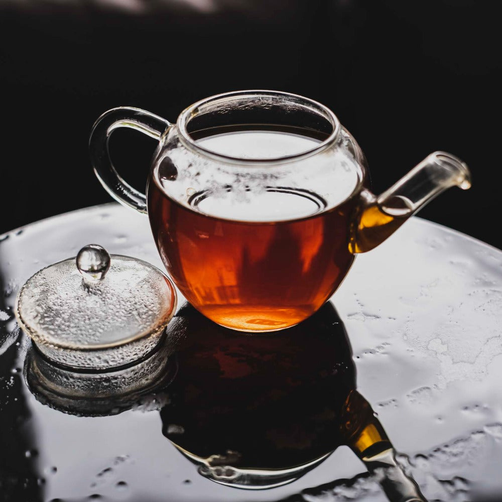 Glasteekanne mit bernsteinfarbenem Tee und abgenommenem Deckel auf schwarzem Untergrund mit Wassertropfen