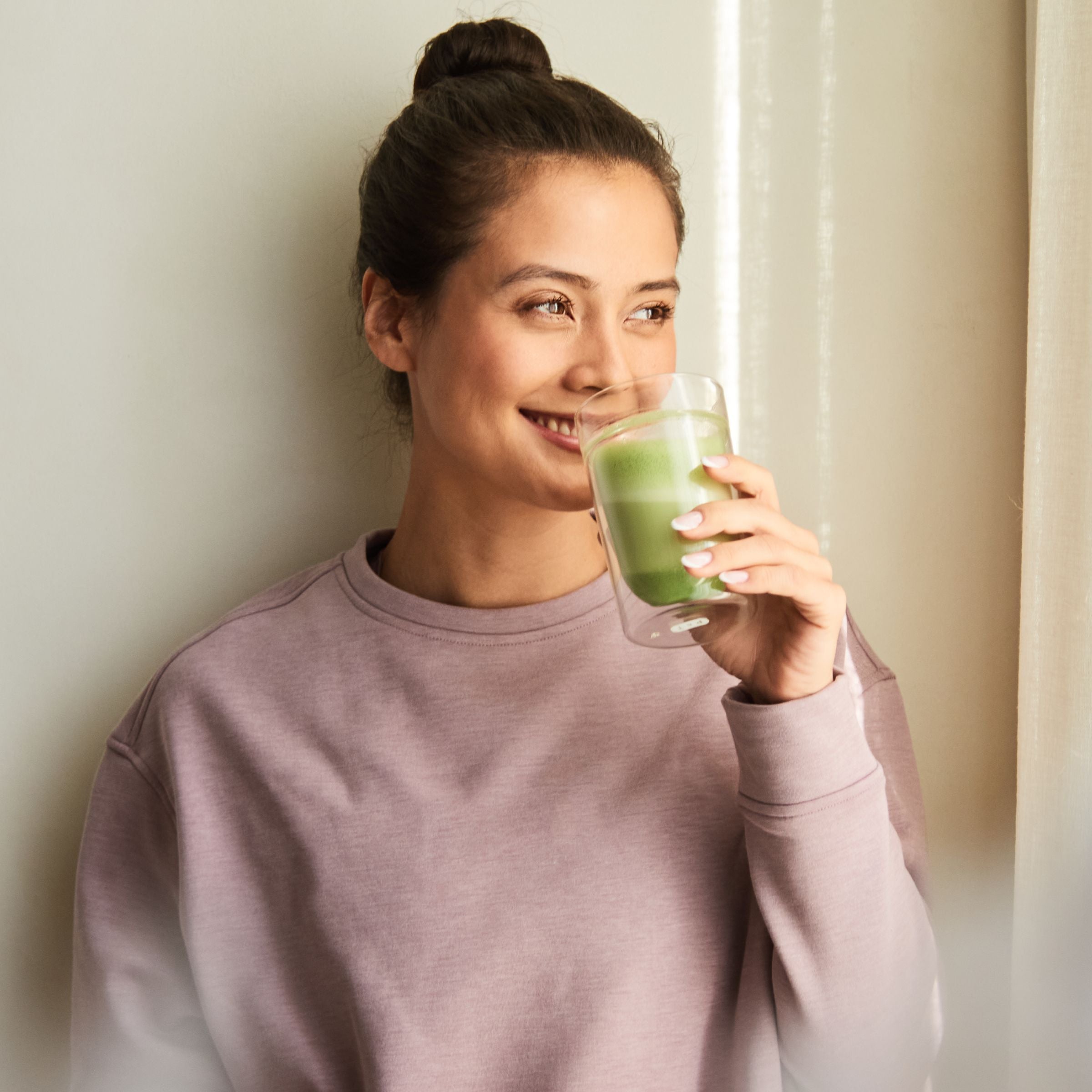 Junge Frau in mauvefarbenem Sweatshirt hält ein doppelwandiges Glas mit grünem Tee und lächelt