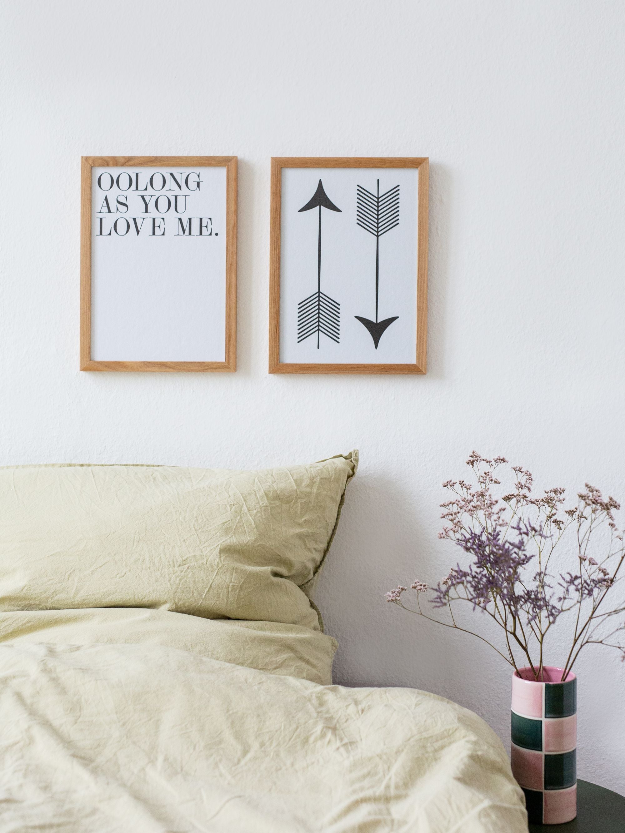 Minimalistisches Schlafzimmer mit Holzrahmen. Links Text: 'OOLONG AS YOU LOVE ME', rechts Pfeile. Beige Bett, schwarzer Tisch mit Vase und lila Blumen.
