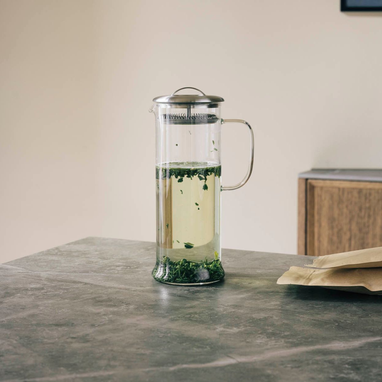 Transparenter Glasteekessel mit Metallsieb, grünem losen Tee neben zwei beigen Papiertüten auf grauer Arbeitsplatte.
