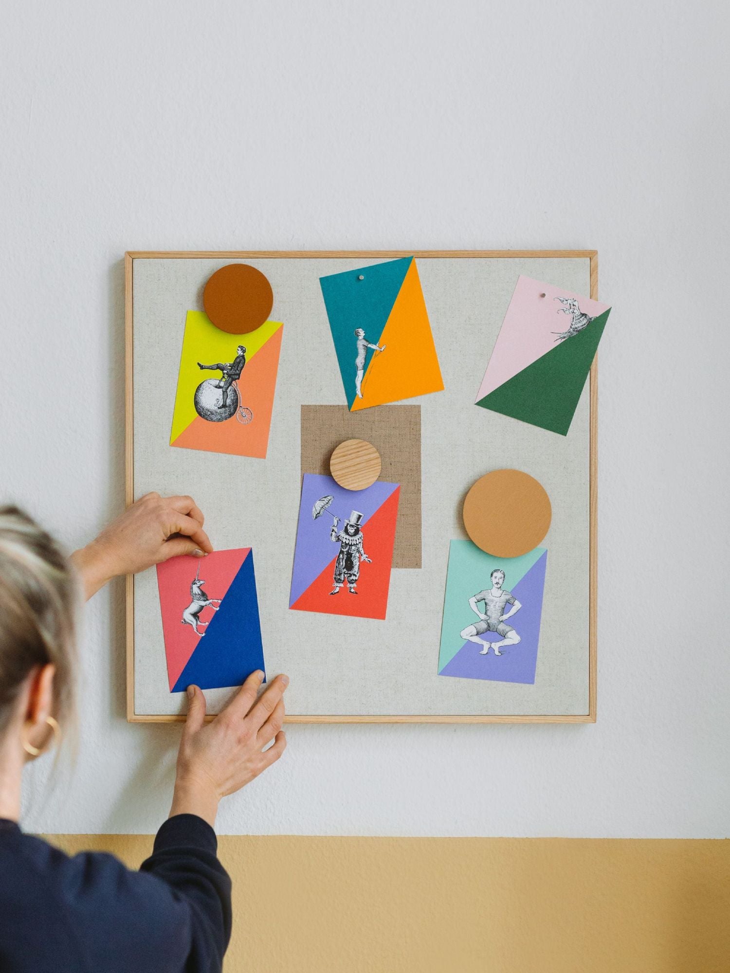Person platziert bunte Vintage-Zirkus-Illustrationen auf einer mit Stoff bezogenen Pinnwand im minimalistischen Raum.