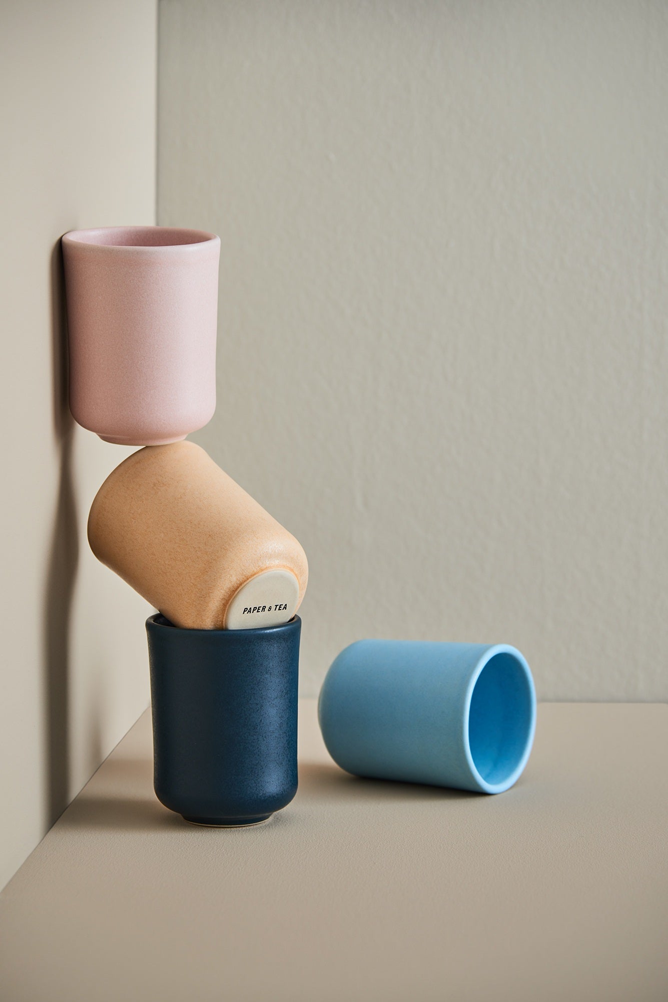 Drei pastellfarbene Keramiktassen, eine mit ‚PAPER & TEA‘ Schriftzug, gestapelt; daneben eine hellblaue Tasse