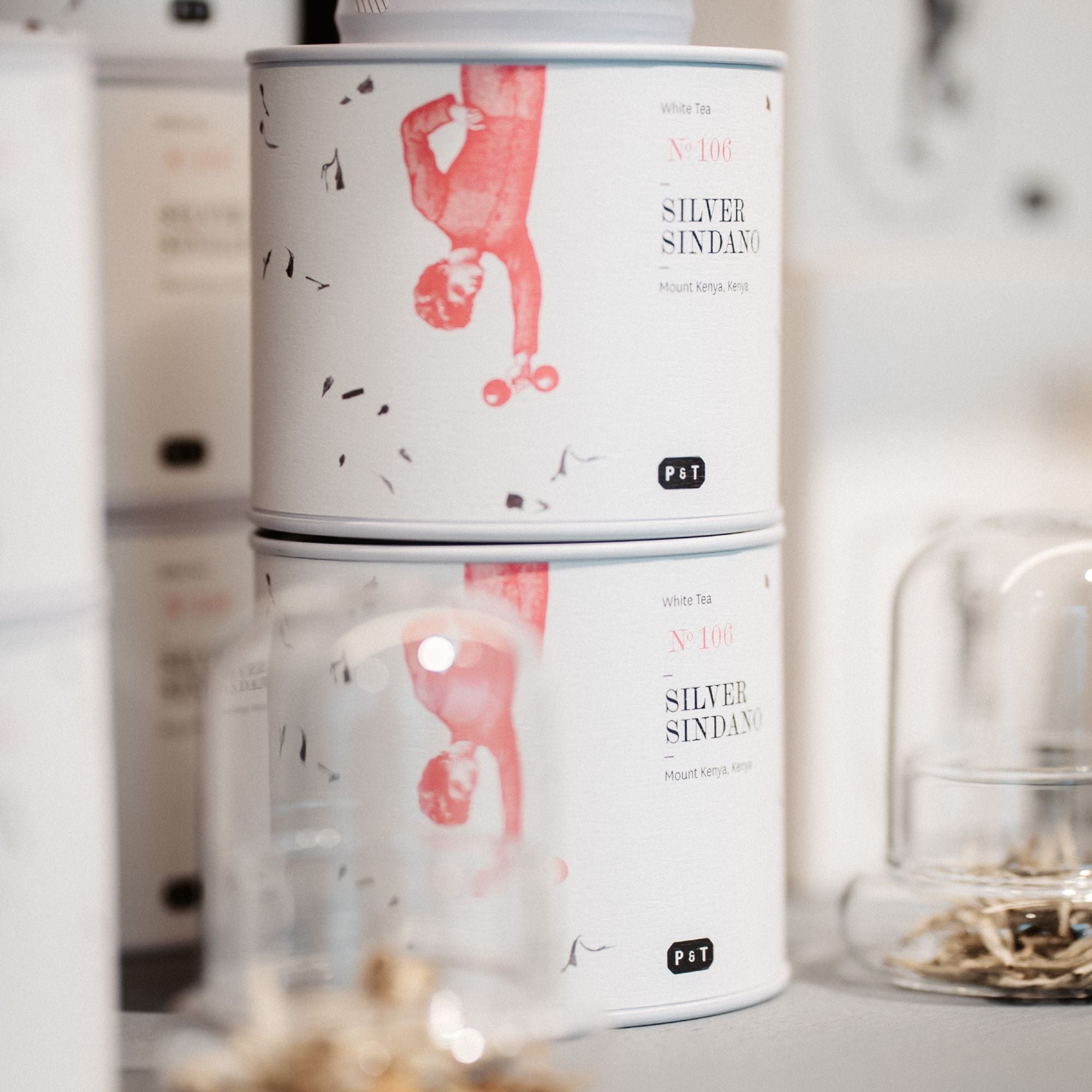 Zwei weiße Teedosen von Paper & Tea mit rotem Silber Sindano-Design, davor Gläser mit Holzdeckeln und Teeblättern.