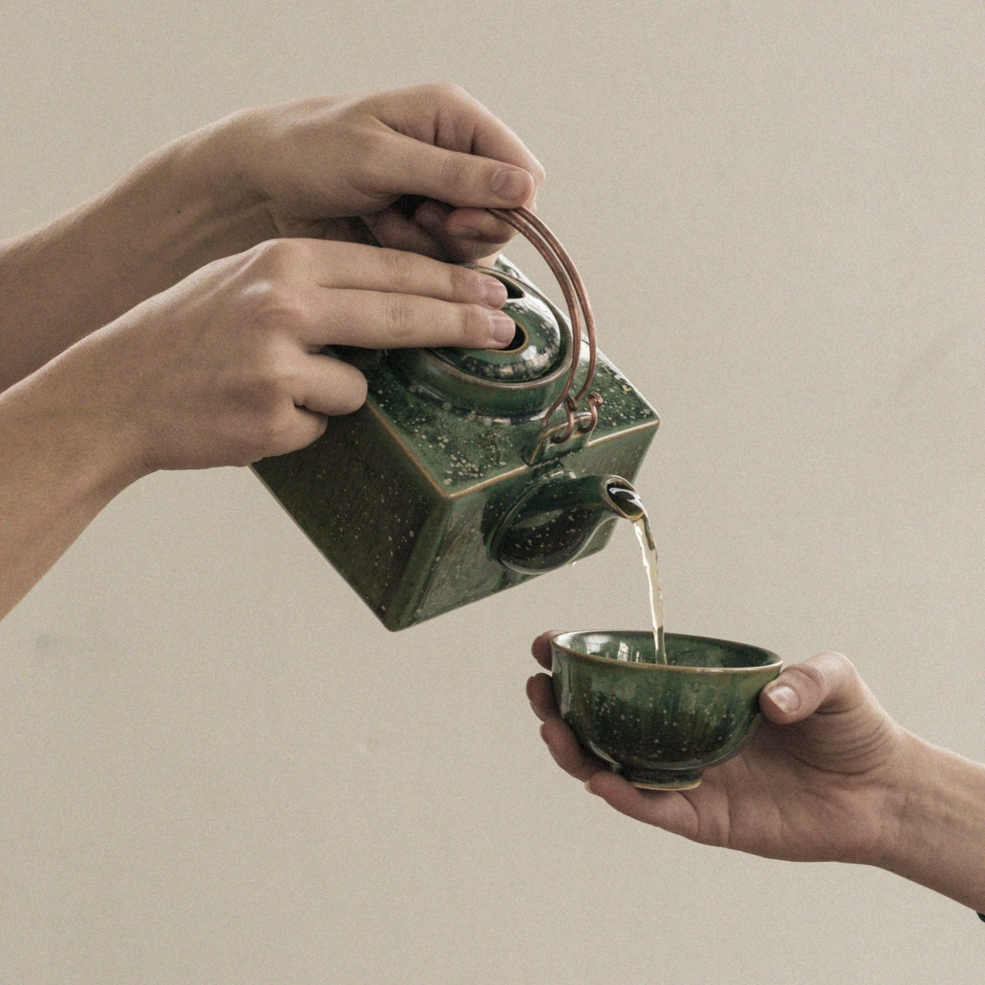 Grüne quadratische Keramik-Teekanne mit braunen Sprenkeln gießt Tee in passenden Becher.