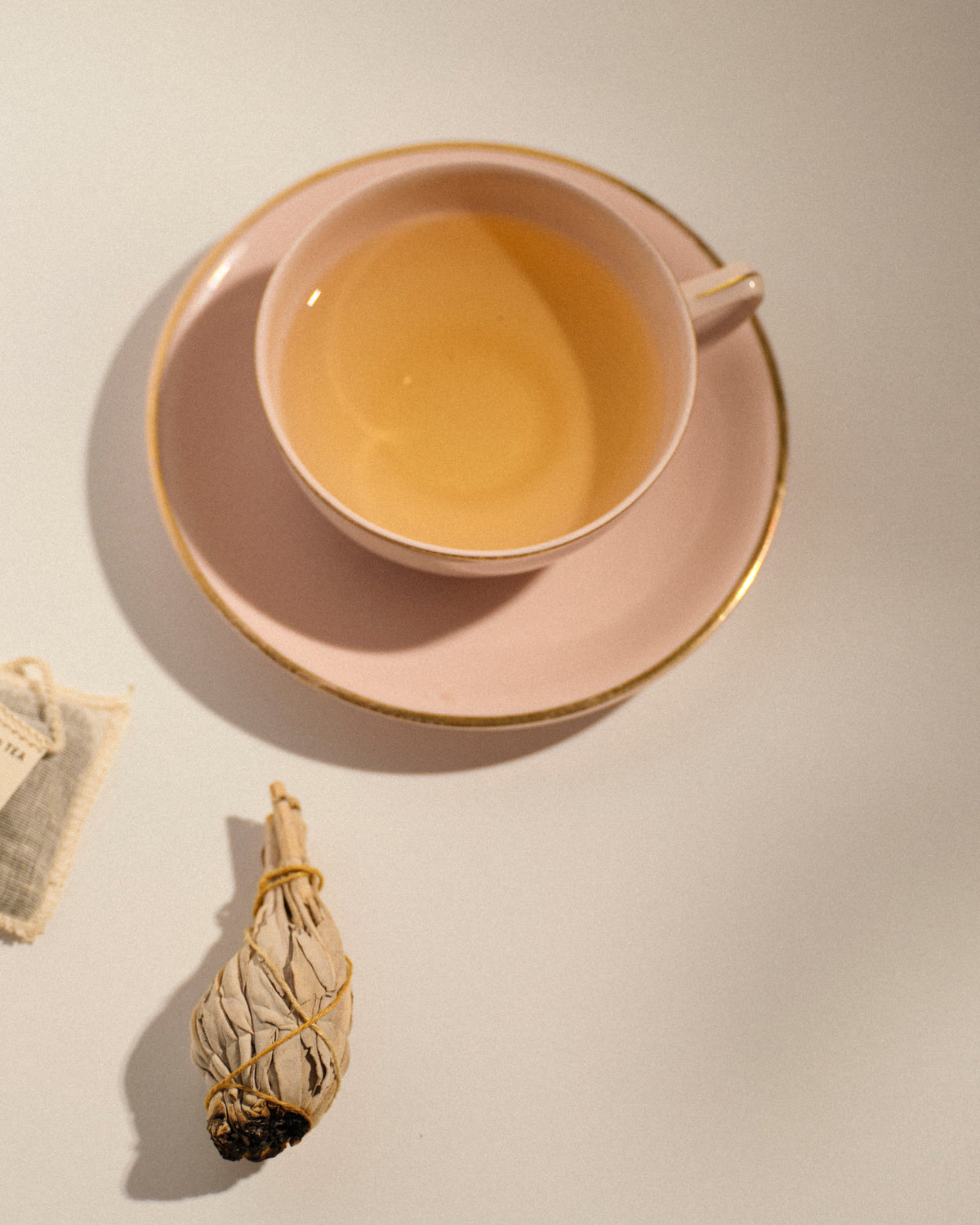 Zartrosa Keramiktasse mit goldgerandetem Unterteller, gefüllt mit hellem Kräutertee, neben gebündelten getrockneten Salbeiblättern und Teebeutel mit Aufdruck P&T.