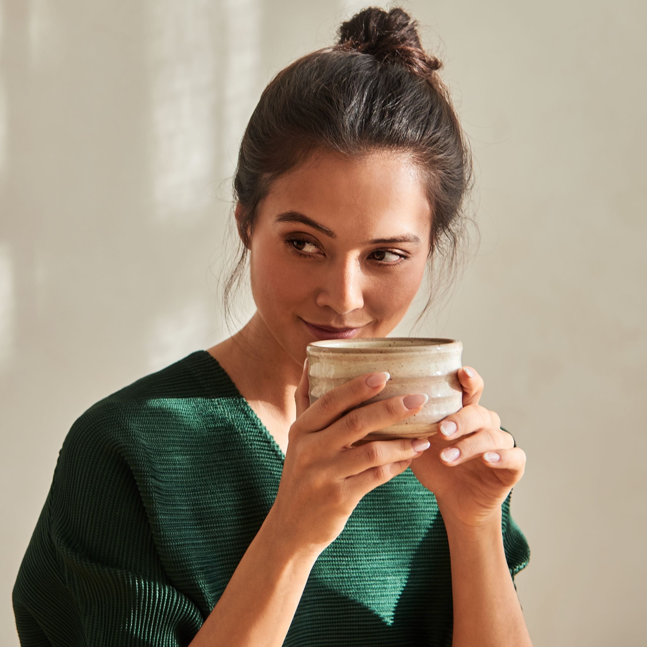 Junge Frau in dunkelgrüner Bluse trinkt Tee aus rustikalem Becher vor warmem, beige beleuchtetem Hintergrund.
