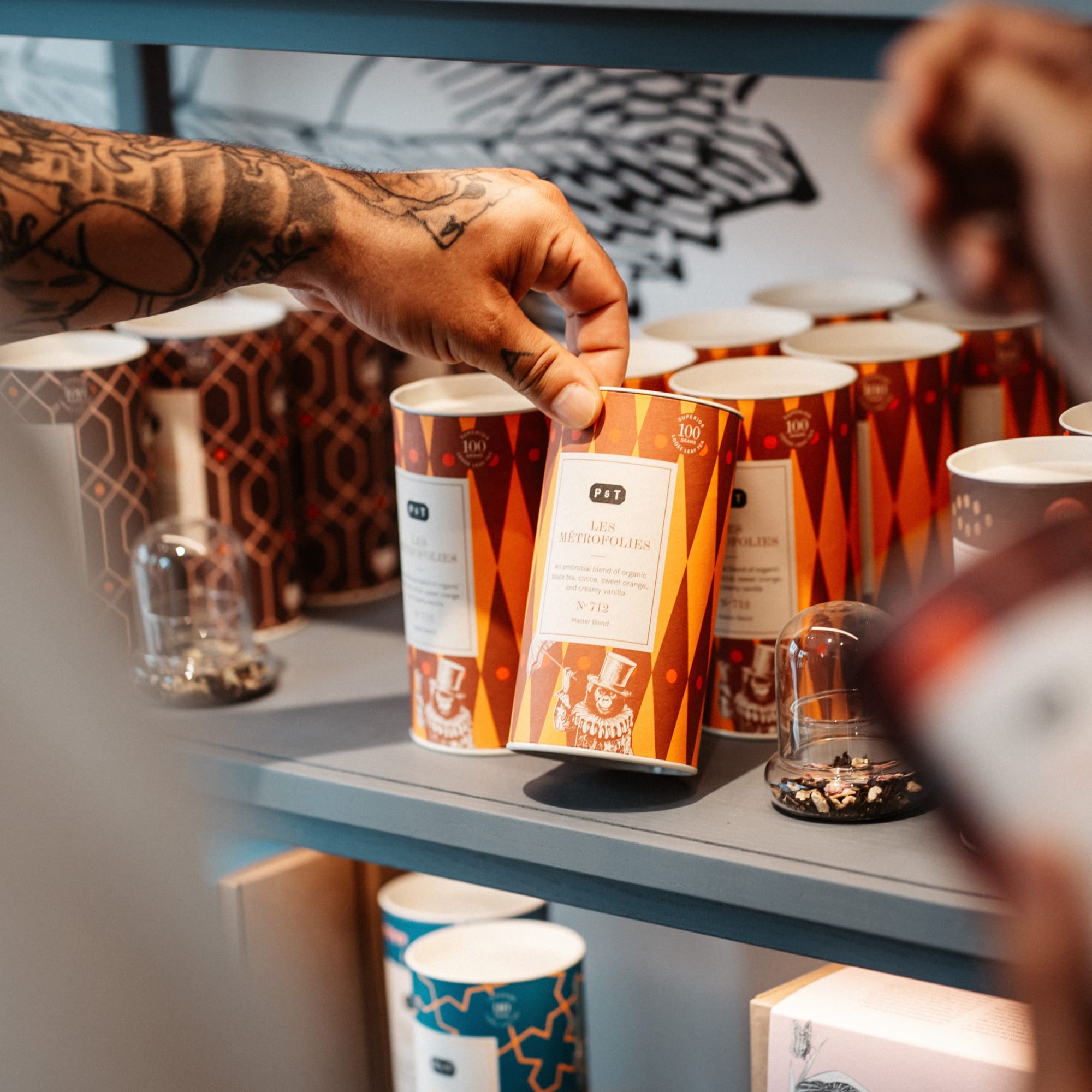 Tätowierte Hand greift nach orange-roter P & T LES MÉTROFOLIES Teedose auf blauem Regal.