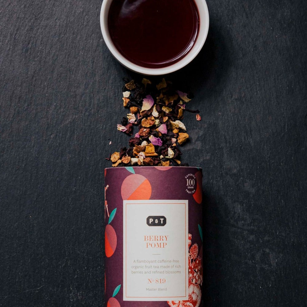 Purpurzylindrische Teedose mit Fruchtillustrationen, lose Bio-Beerenteemischung und weiße Tasse auf dunklem Hintergrund.