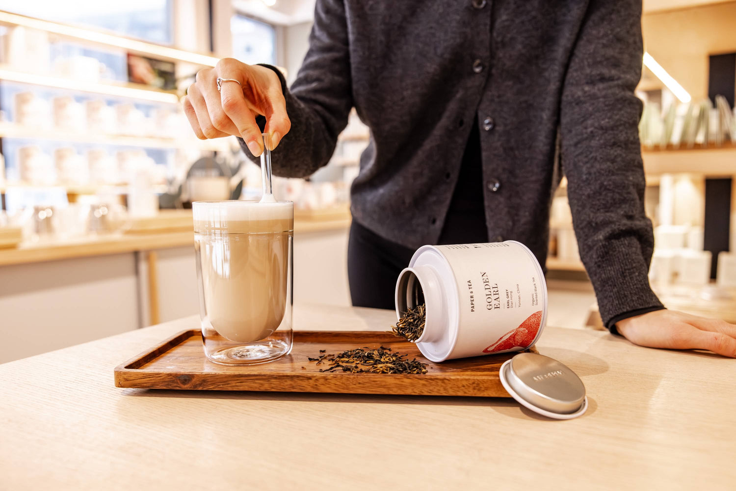 Person rührt in einem Glas schaumigem Earl Grey Getränk neben einer geöffneten Paper & Tea Teedose