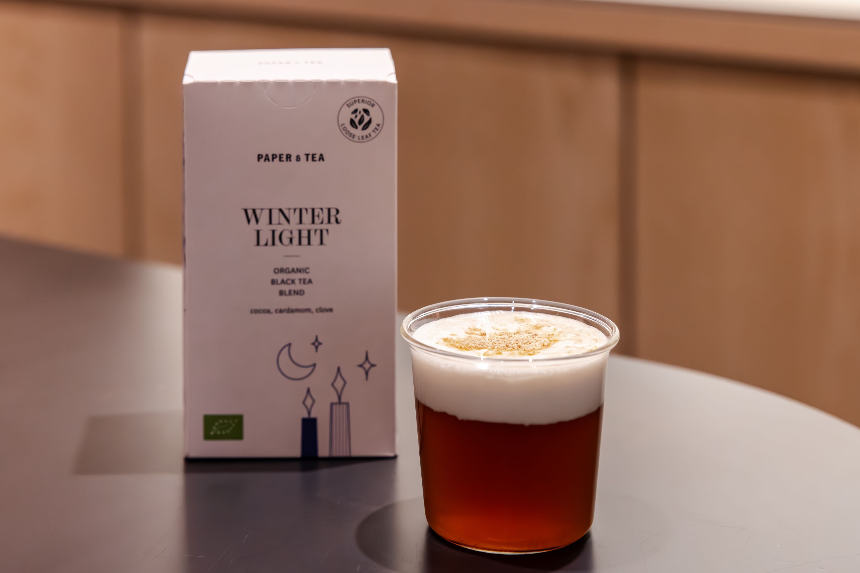 Weiße Box mit Winter Light Bio-Schwarztee neben klarem Becher mit schaumigem, gewürztem schwarzen Tee