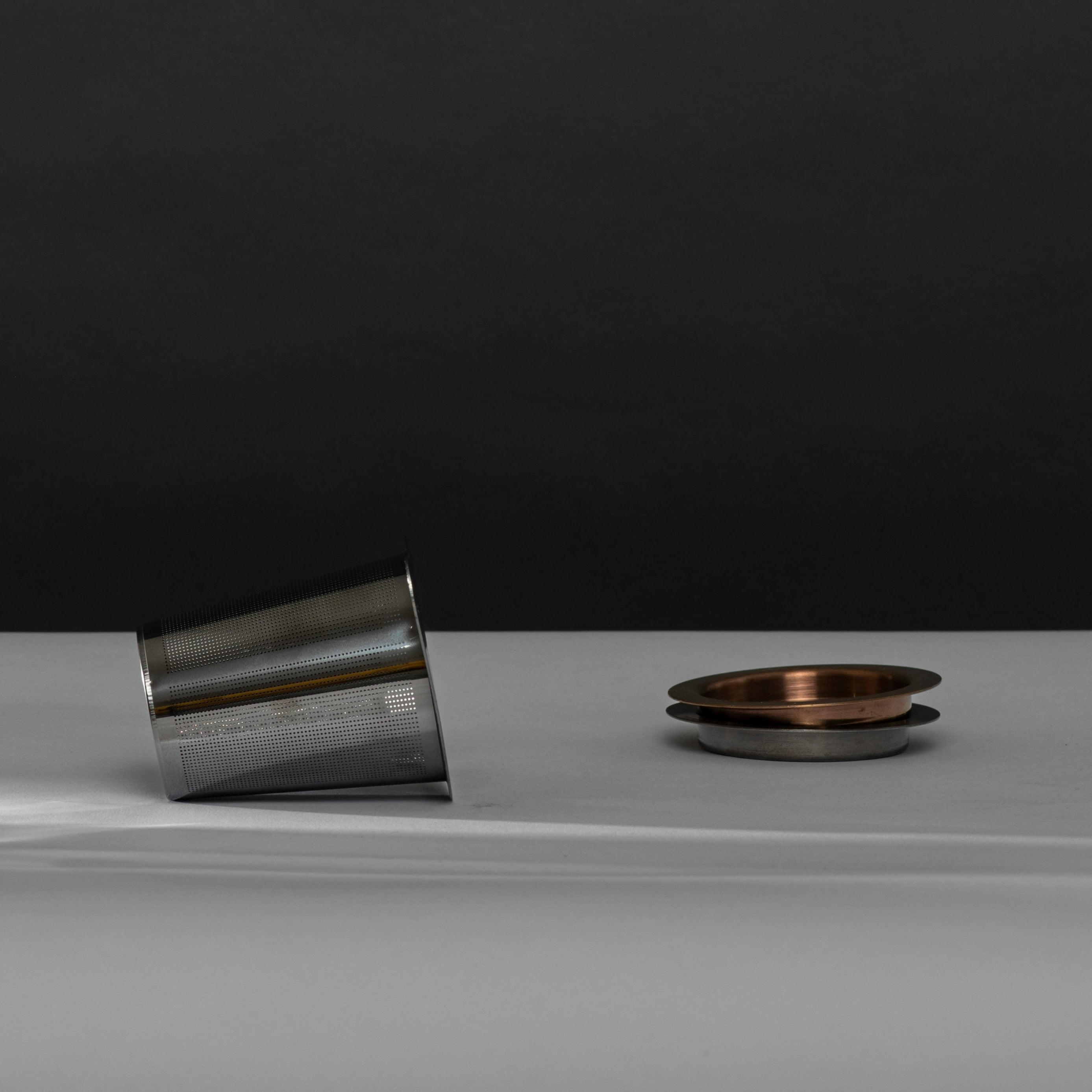 Metallischer Teesieb mit feinen Löchern neben gestapelten Kupfer- und Silberschalen auf hellgrauem Tisch