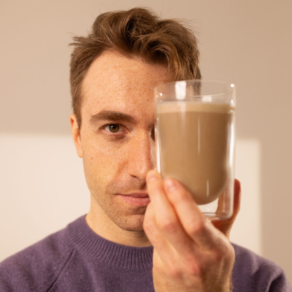 Ein Mann mit braunen Haaren hält ein Glas mit cremigem, hellbraunem Getränk nah an seinem Gesicht.