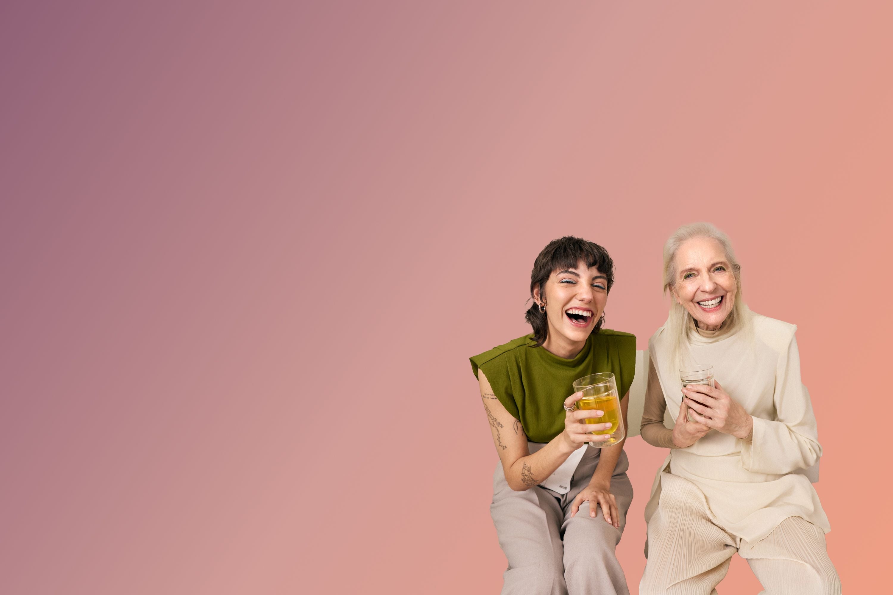 Zwei Frauen, eine mit olivgrünem Oberteil und Tattoos, lachen und halten Teegläser vor buntem Hintergrund.