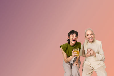 Zwei Frauen, eine mit olivgrünem Oberteil und Tattoos, lachen und halten Teegläser vor buntem Hintergrund.
