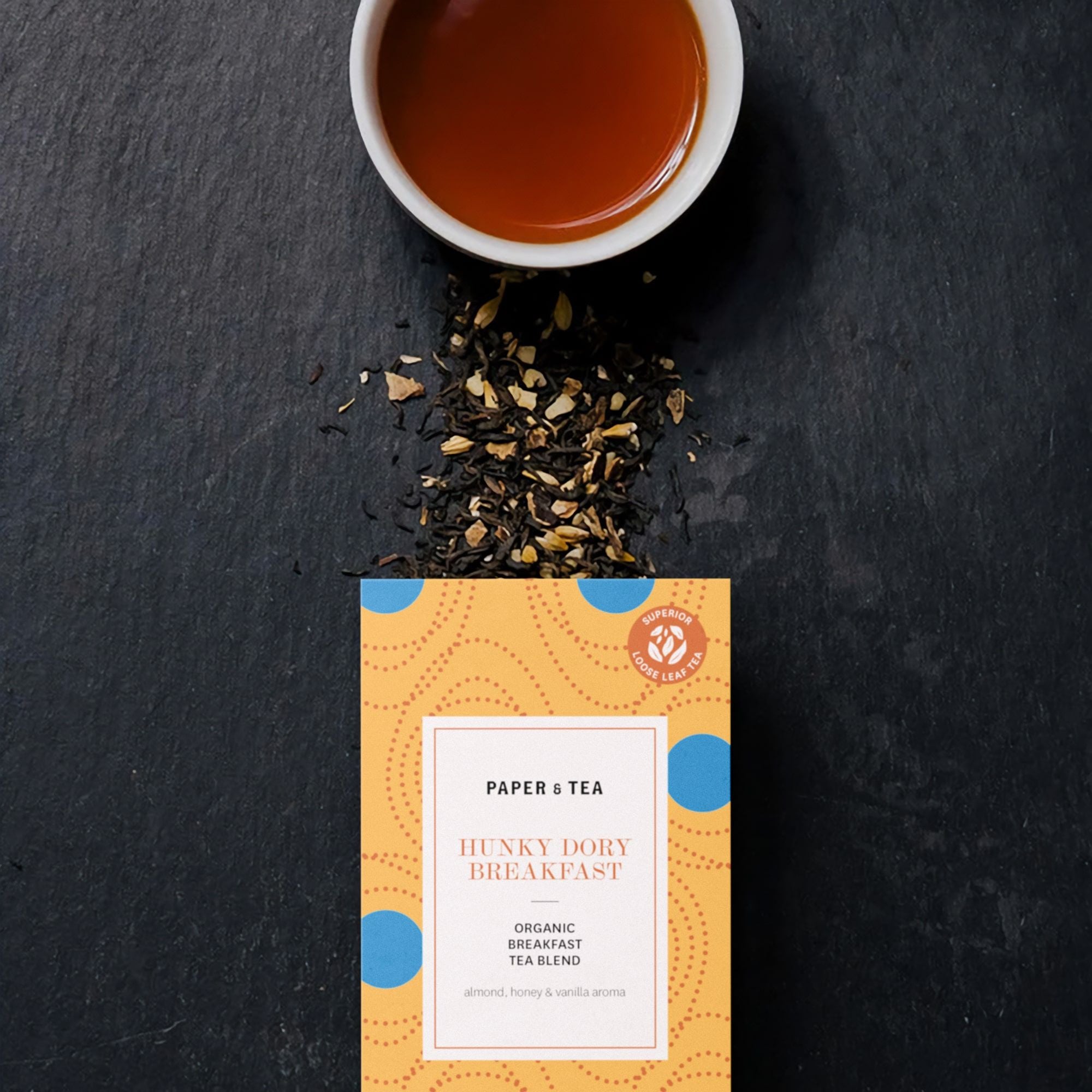 Weiße Tasse mit Hunky Dory Breakfast Bio-Tee auf Schiefer, orange PAPER & TEA Verpackung daneben.
