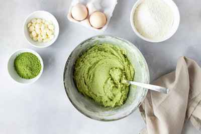 Metallschüssel mit grünem Matcha-Teig und Zutaten wie Eier, weiße Schokoladenstückchen, Matcha-Pulver und Kuchenmehl.