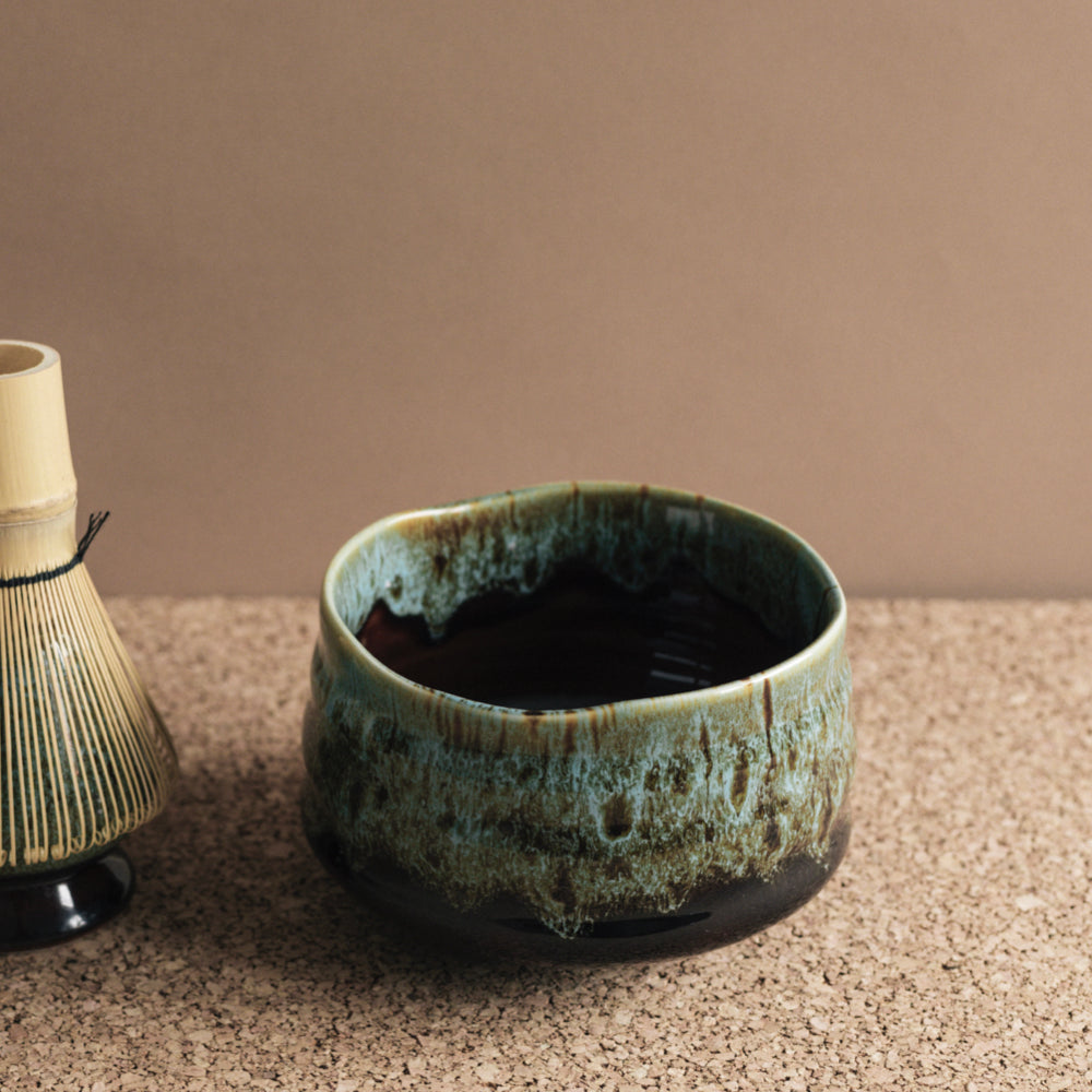 Keramische Teeschale mit unregelmäßigem Rand und dunklem Tee neben traditionellem Bambusbesen auf Korkuntergrund.