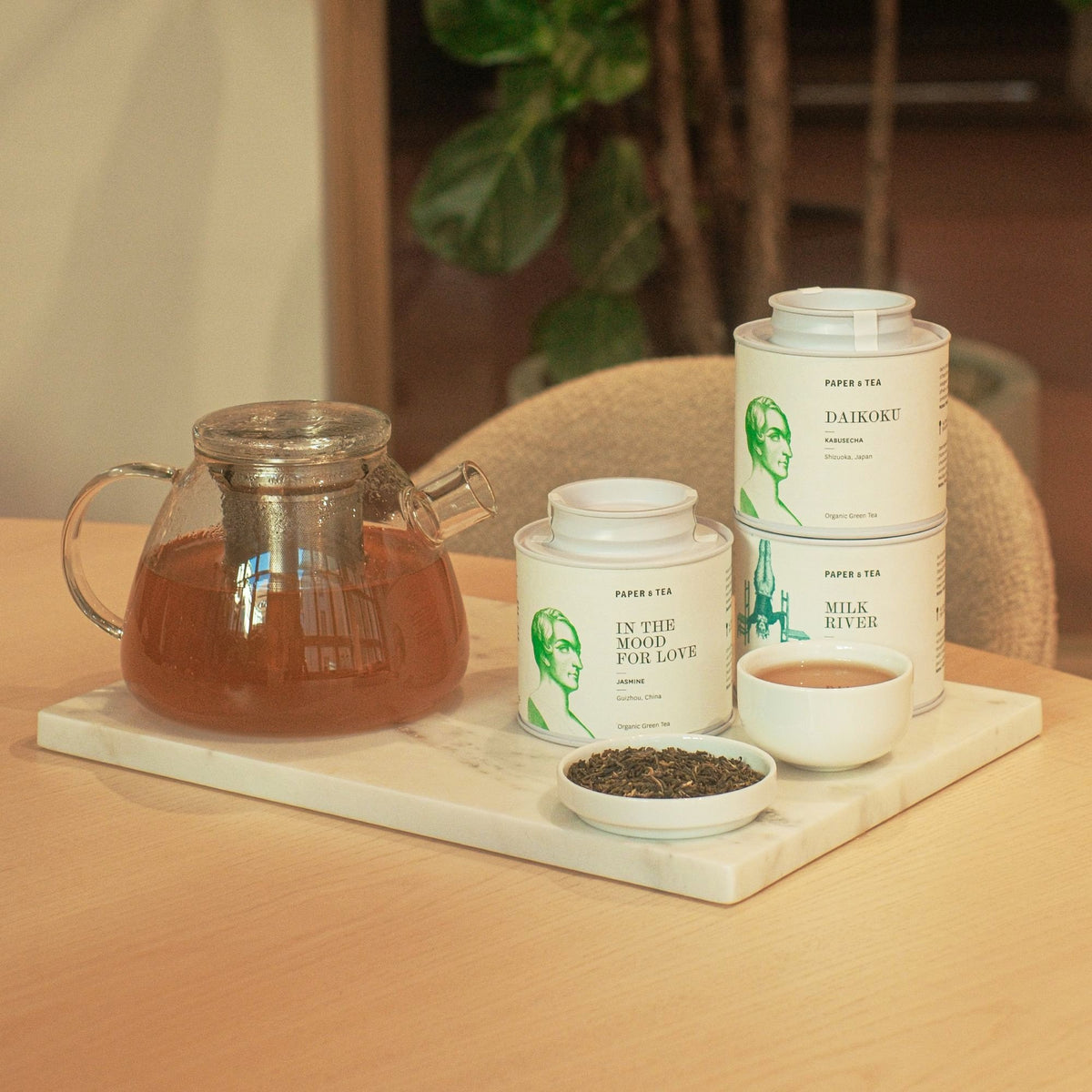 Weißes Marmortablett mit Glasteekanne, drei Teedosen und grünen Teeblättern.