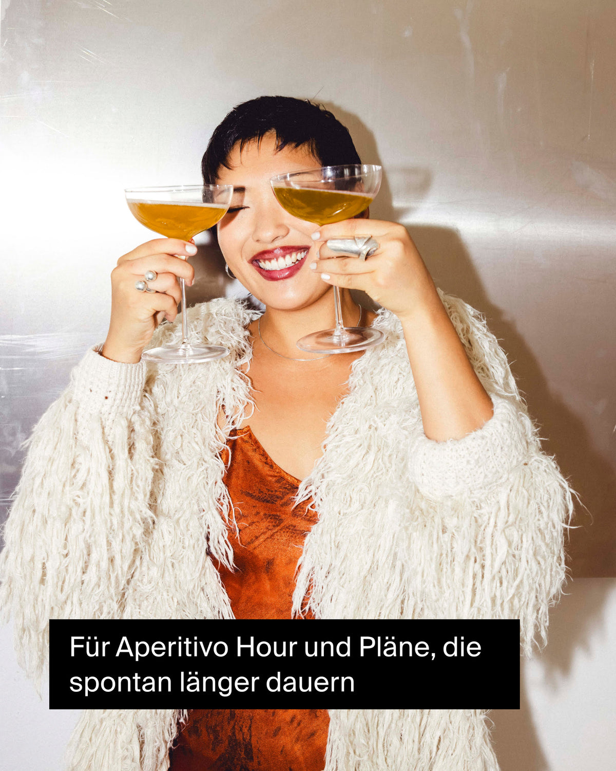 Fröhliche Frau mit kurzen schwarzen Haaren in weißer Jacke, hält zwei goldene Cocktails zum Aperitivo.