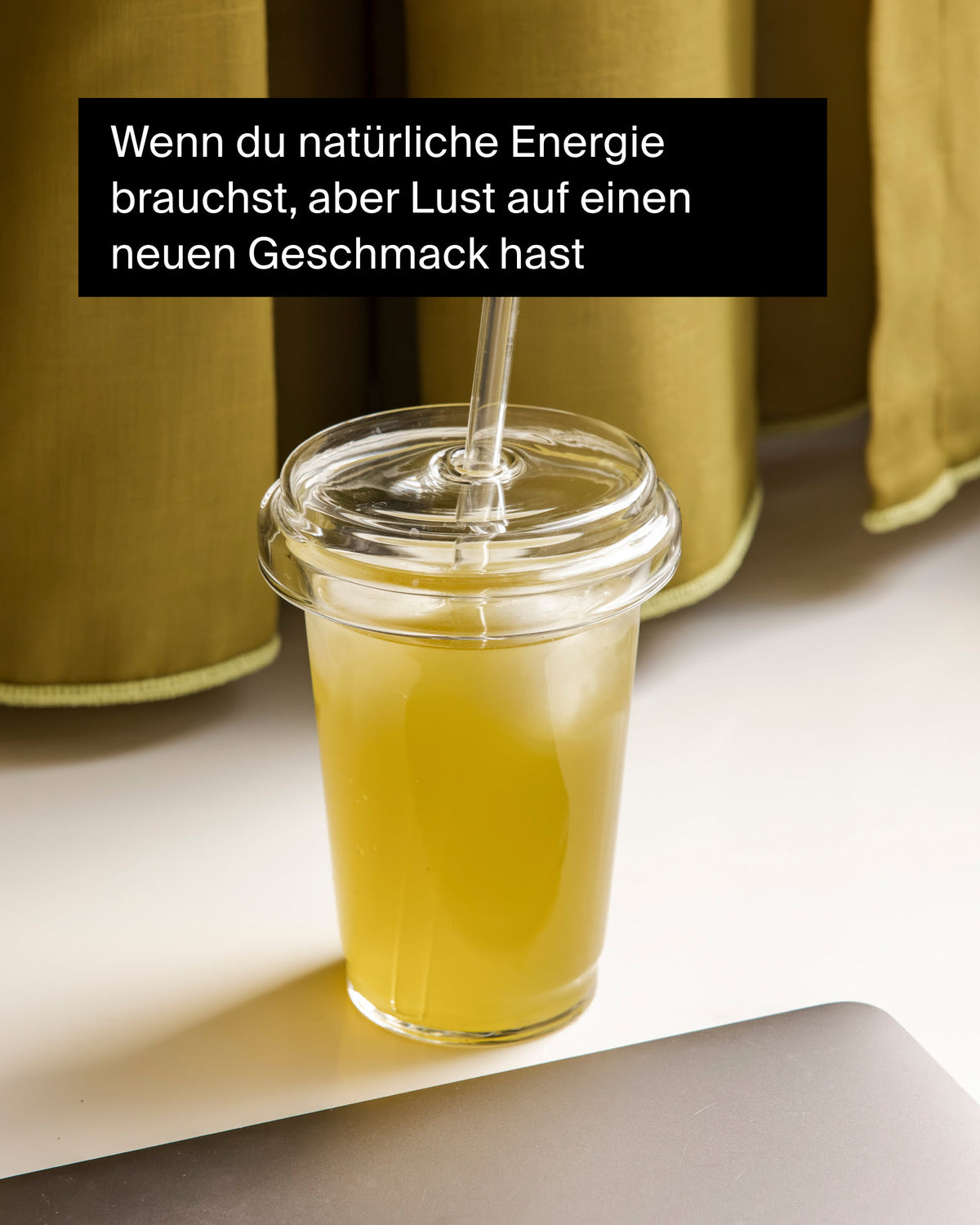 Transparenter Becher mit gelb-grünem Eistee und Eiswürfeln auf weißer Fläche, daneben ein silbernes Laptop