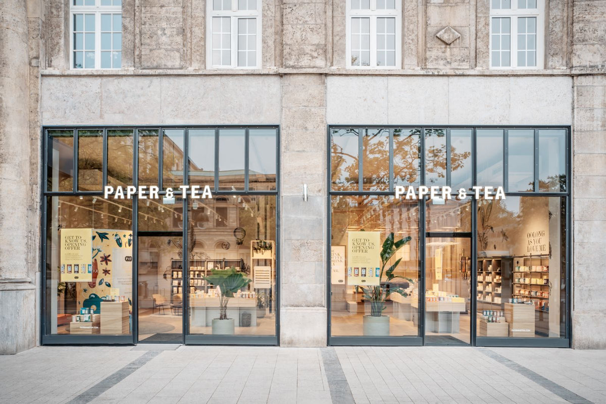 Außenansicht des P&T Teeladens mit großen Schaufenstern, innen Teeprodukte, Pflanzen und Werbeplakate.