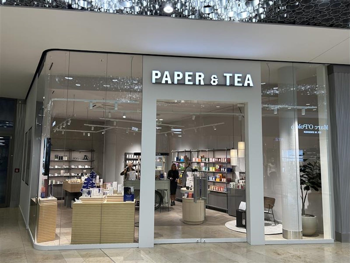 Moderner PAPER & TEA Laden mit Glasfront, bunte Teeprodukte im Schaufenster, zwei Frauen im Gespräch.