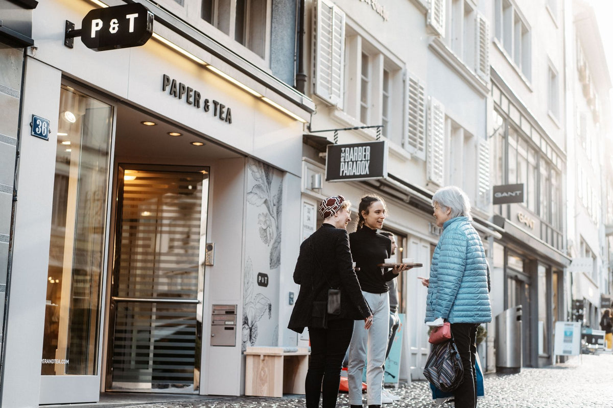 Eingang des modernen PAPER & TEA Ladens mit schwarzem P & T Schild, drei Frauen beim Tee auf Kopfsteinpflaster