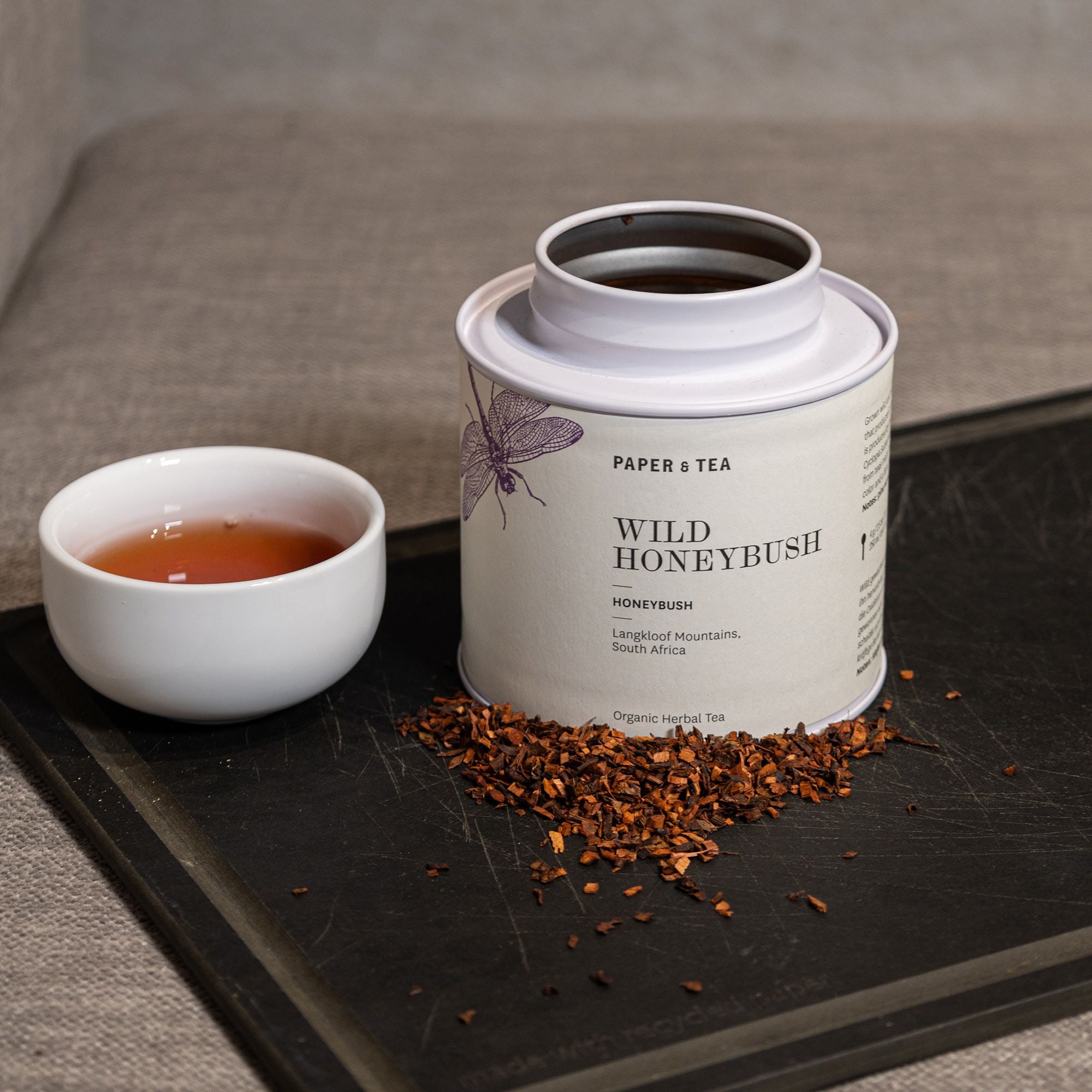 Weißer Zylinderbehälter mit WILD HONEYBUSH Bio-Kräutertee von PAPER & TEA auf schwarzem Tablett.